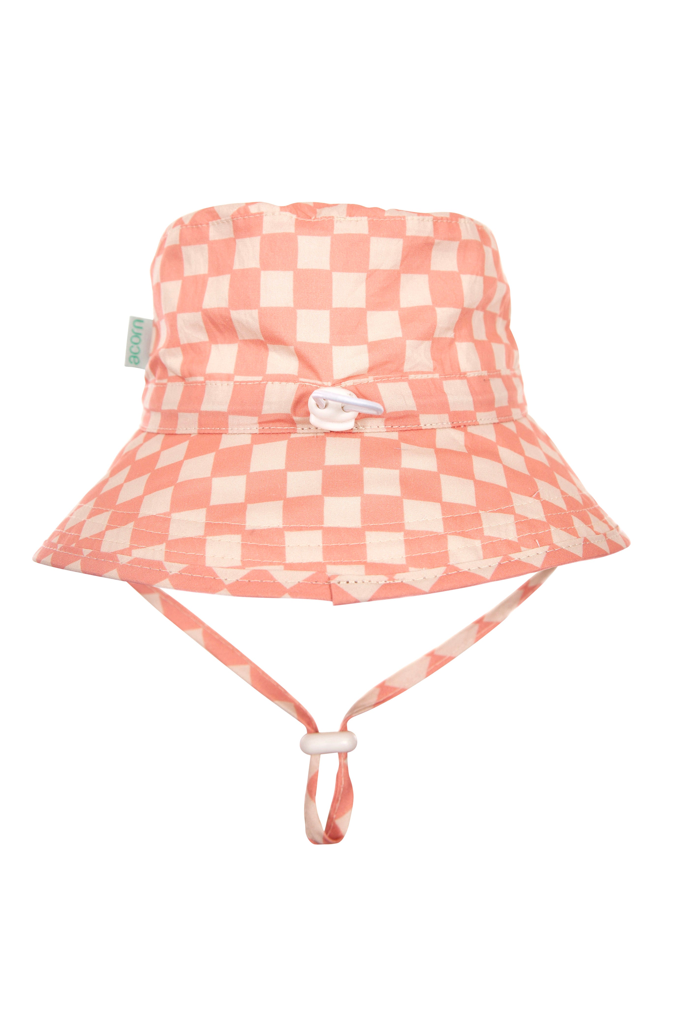 The Pink Checks Broad Brim Bucket Hat by ACORN KIDS is crafted from 100% cotton for optimal sun protection and features a pink and white checkered pattern, a chin strap, and a small tag on the side.