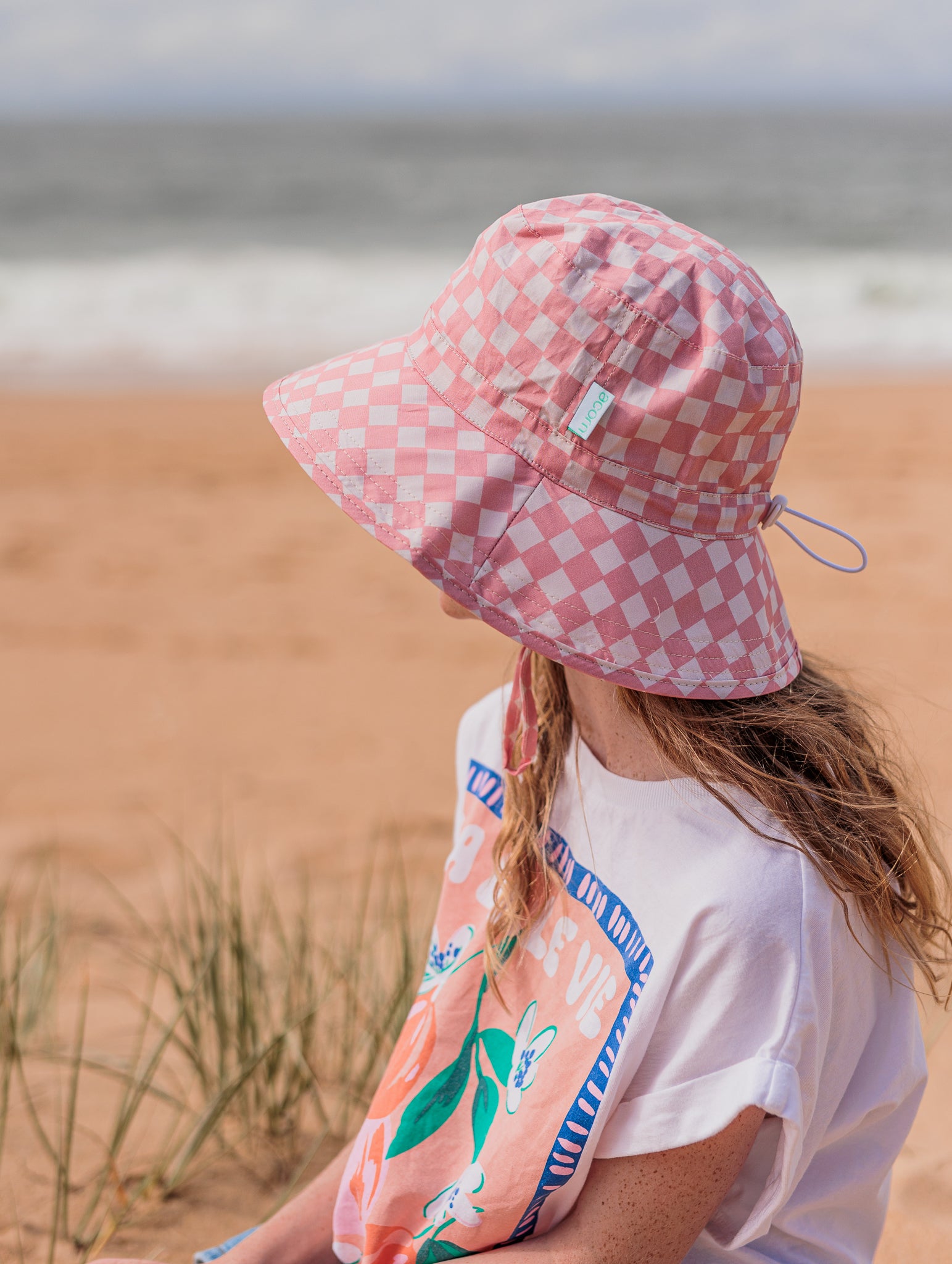 A woman sitting on a sandy beach wears the Pink Checks Broad Brim Bucket Hat from ACORN KIDS, crafted from 100% cotton. She pairs it with a white t-shirt showcasing colorful graphics. Ocean waves crash gently in the background under a cloudy sky, while patches of grass dot the sand, providing her with stylish sun protection.