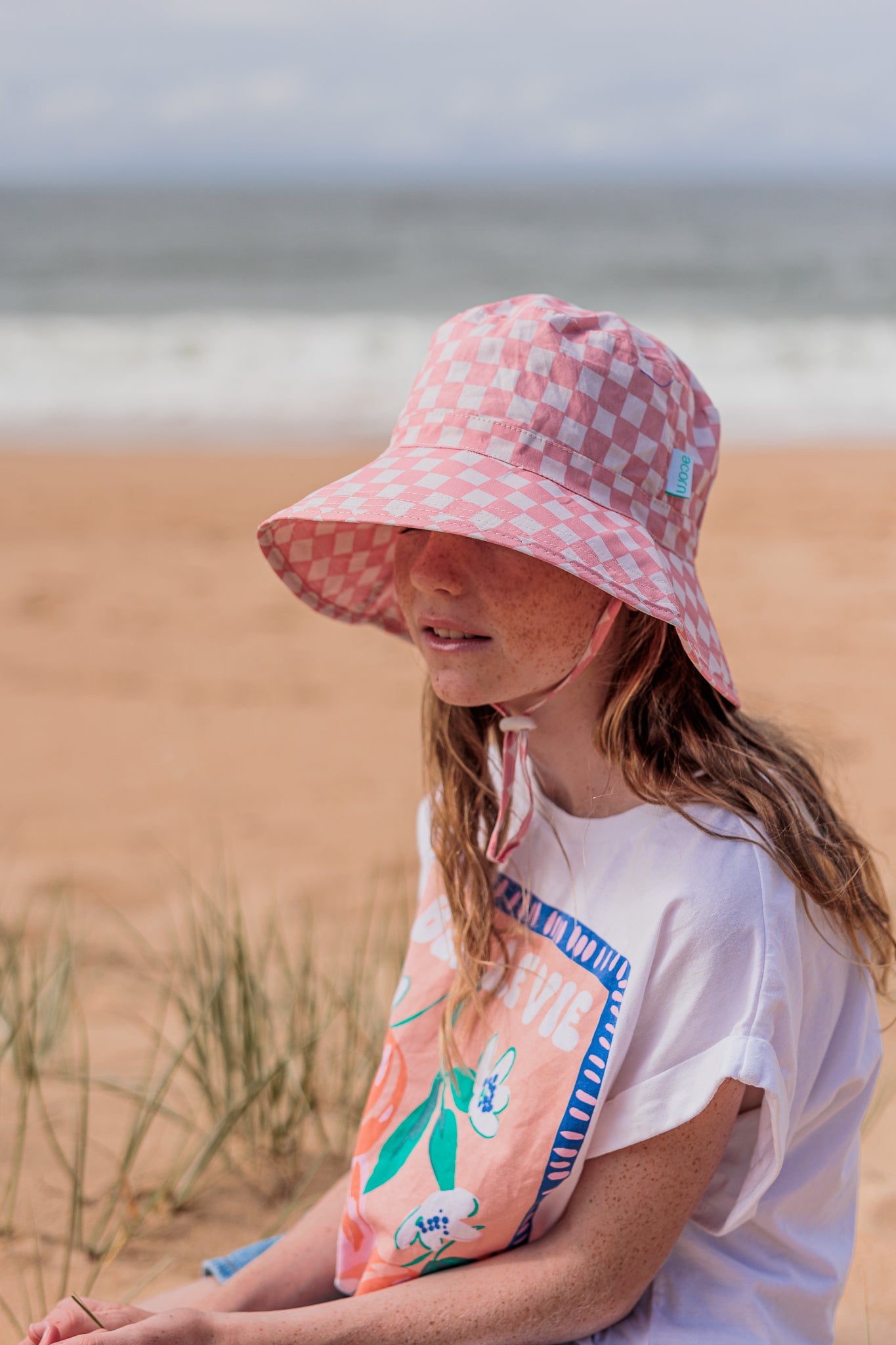 A person wearing the ACORN KIDS Pink Checks Broad Brim Bucket Hat that offers UPF50+ sun protection and a white T-shirt with a colorful design sits on the beach near the sea. Grass peeks through the sand in the foreground, with the ocean and sky stretching out in the background.