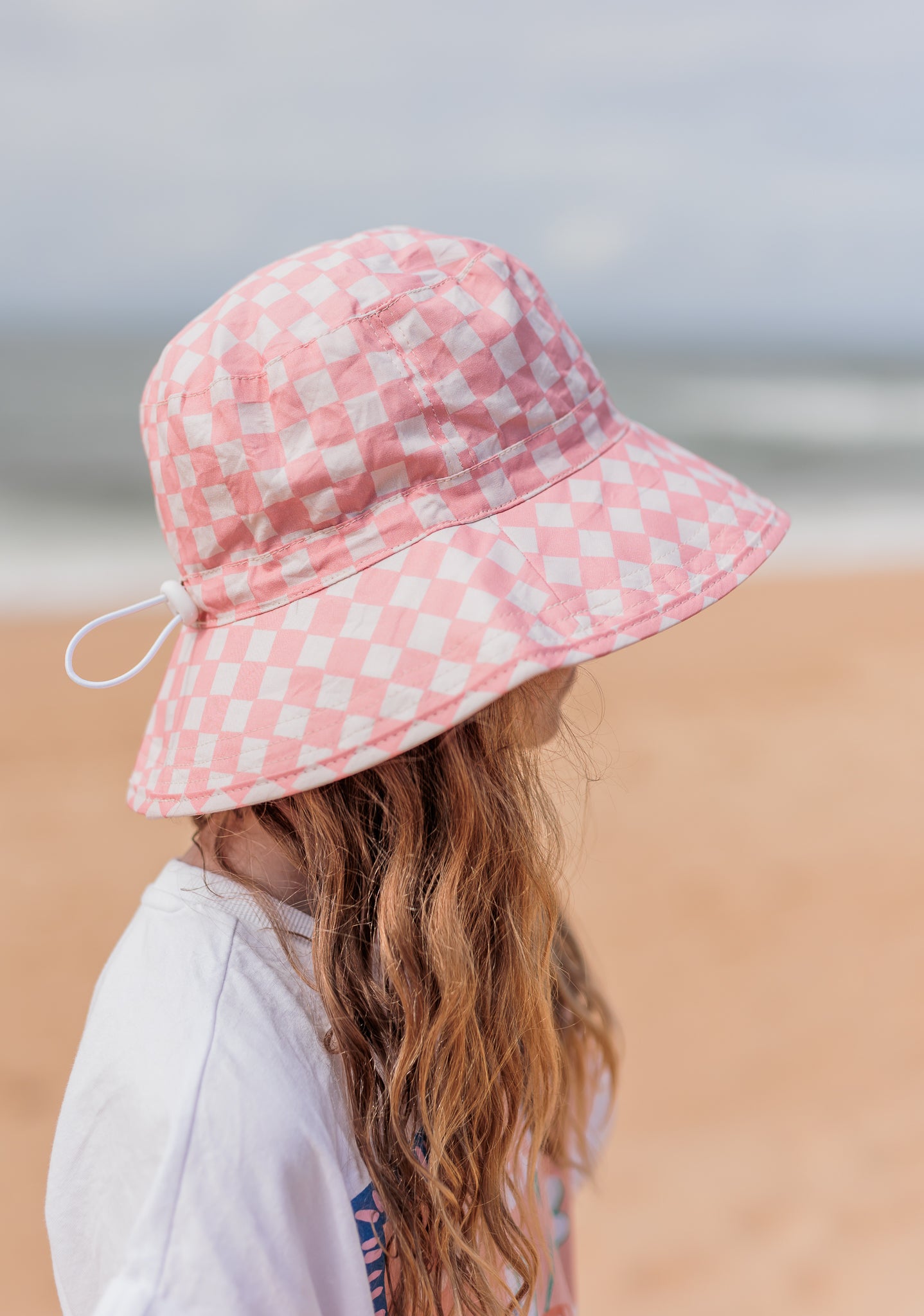 A person with long hair is standing on a beach, wearing the Pink Checks Broad Brim Bucket Hat by ACORN KIDS. The ocean and sandy shore are blurred in the background, creating a serene scene. The UPF50+ sun protection ensures they’re well-shielded as they turn slightly away from the camera.