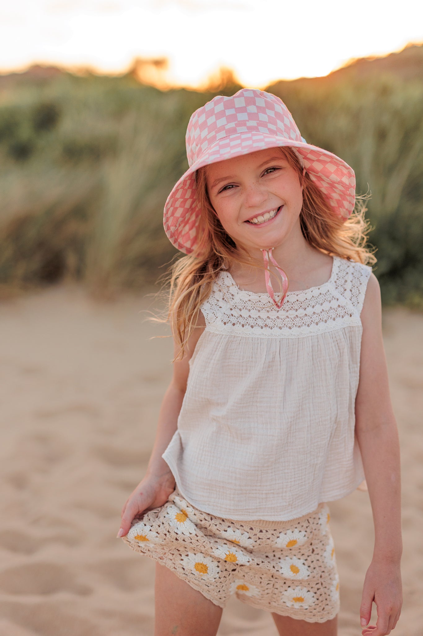 A young girl smiles on a sandy beach at sunset, wearing the ACORN KIDS Pink Checks Broad Brim Bucket Hat with UPF50+ sun protection, a white sleeveless top, and floral-patterned shorts. The background showcases grassy dunes and a glowing sky.