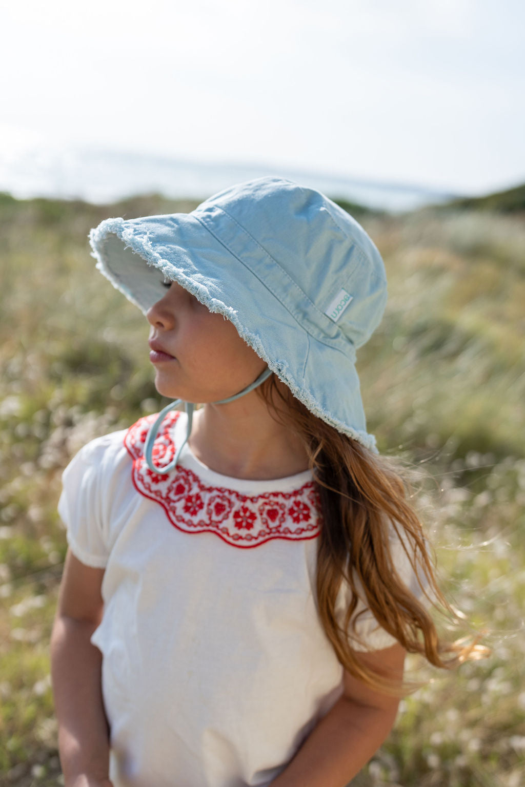 A young girl with long hair stands outdoors, wearing the ACORN KIDS Frayed Bucket Hat in Seafoam, which offers UPF50+ sun protection. She is also dressed in a white shirt featuring red embroidery around the neckline. As she gazes to the side, grassy fields and a blurry ocean serve as her backdrop.