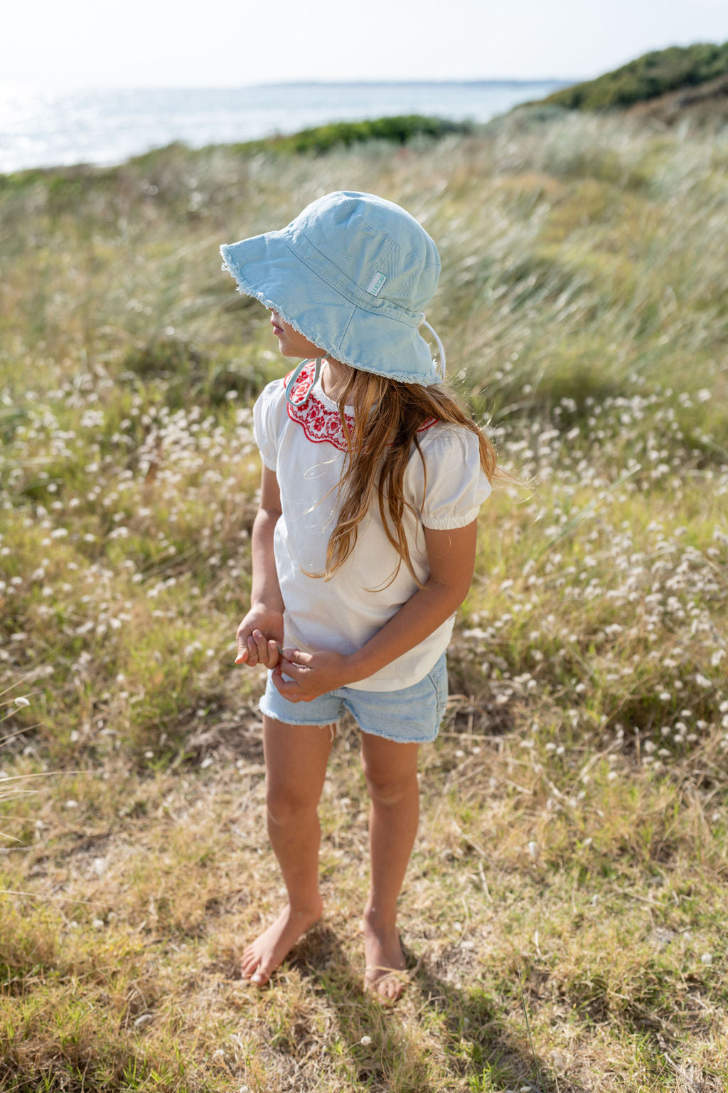 A young girl stands in a grassy field near the sea, looking to her right. She wears a Frayed Bucket Hat Seafoam from ACORN KIDS, featuring UPF50+ sun protection, a white T-shirt with a red embroidered neckline, and light blue denim shorts. The wind gently blows her hair and the field's tall grass, with the sea visible in the background.