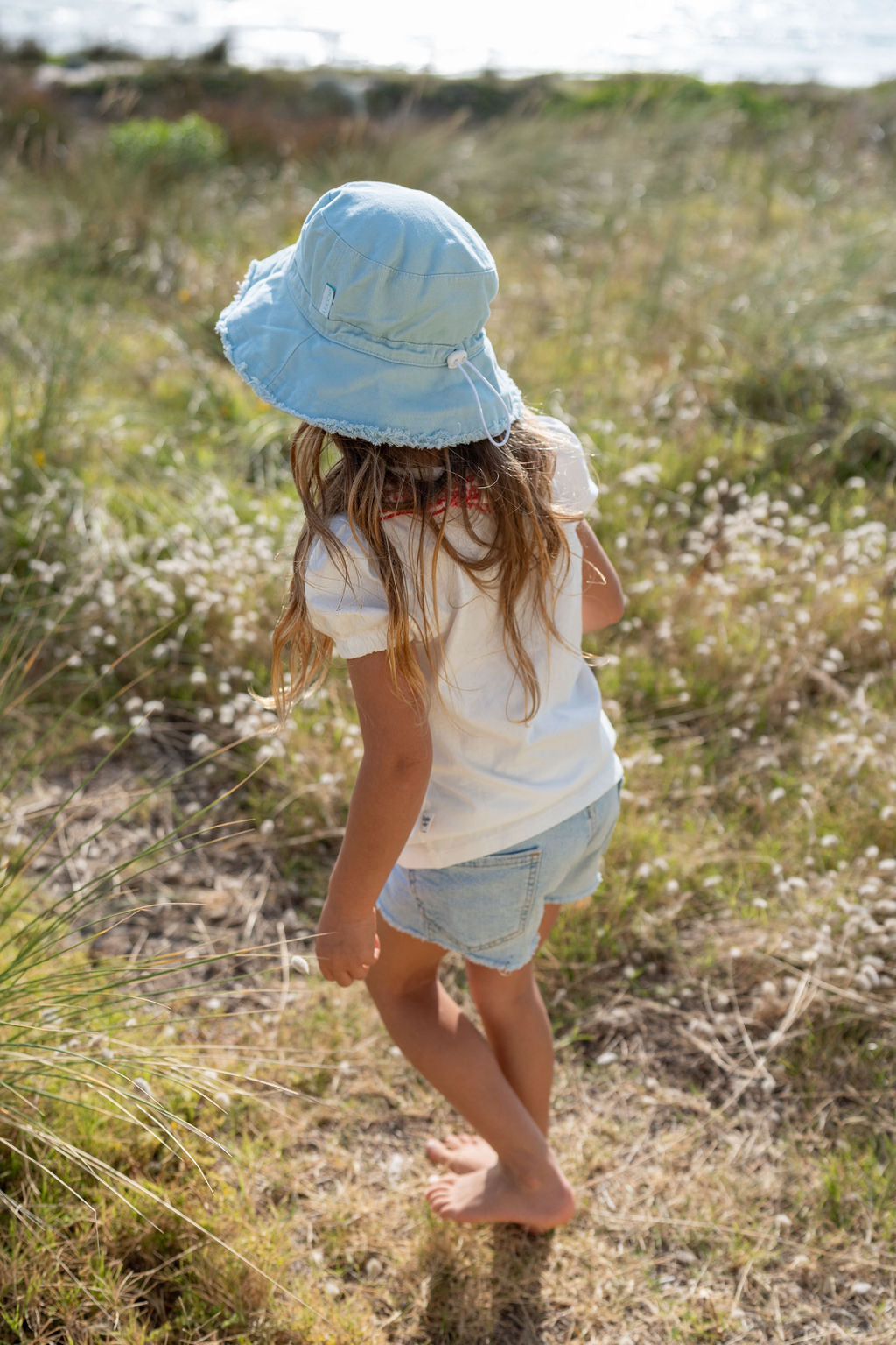 A young child with long hair, wearing the ACORN KIDS Frayed Bucket Hat Seafoam for UPF50+ sun protection, a white t-shirt, and denim shorts, walks barefoot through a grassy field dotted with wildflowers. The scene is bathed in sunlight, and a body of water is visible in the background.