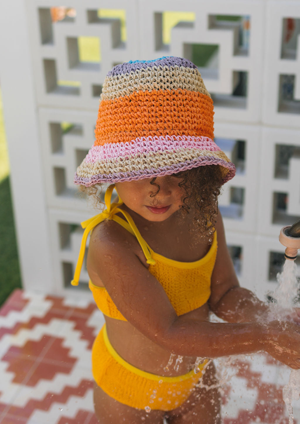 A young child in a yellow swimsuit wears the ACORN KIDS Acorn Kids - Astrid Crochet Straw Hat while playing with water from a faucet, standing on patterned tiles in front of a decorative white wall.