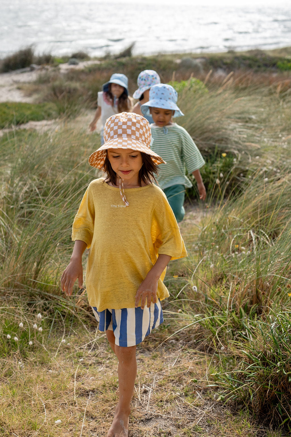 Four children walk along a coastal path with sandy ground and greenery. The first child wears a yellow shirt, blue and white striped shorts, and an ACORN KIDS Checkmate Wide Brim Bucket Hat with caramel and cream checks. The other children follow behind, also wearing sun hats offering UPF50+ sun protection and casual summer clothes. The sea is visible in the background.