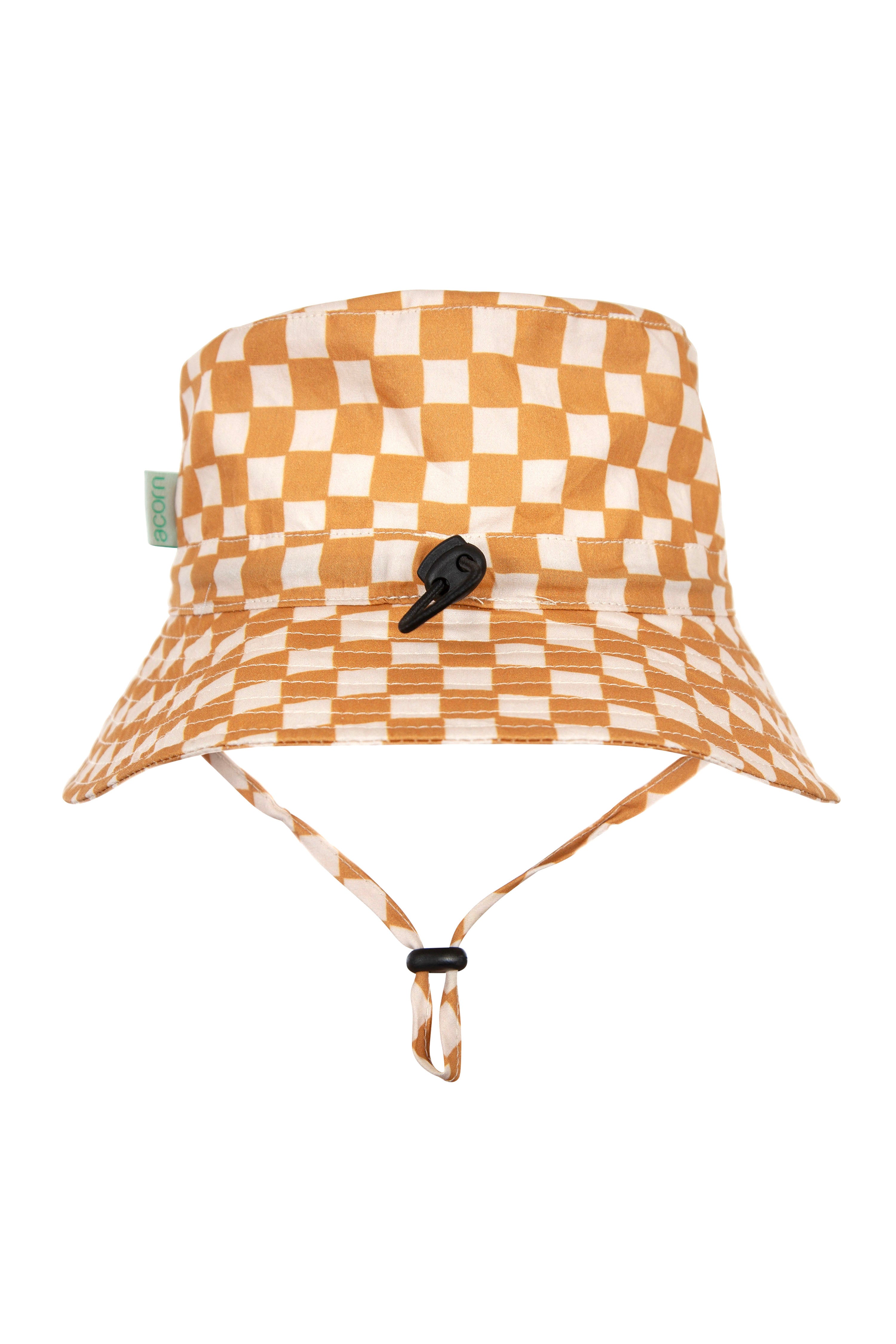 The Checkmate Wide Brim Bucket Hat by ACORN KIDS features caramel and cream checks, offers UPF50+ sun protection, includes a green tag on the side, has an adjustable black cord around the brim, and comes with a drawstring chin strap with a black toggle.