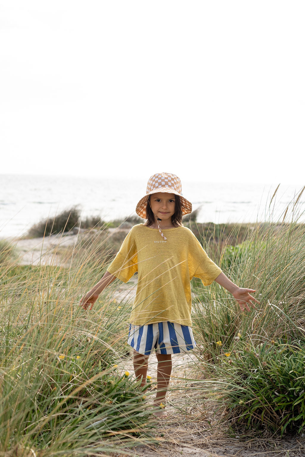 A child wearing a yellow t-shirt, blue and white striped shorts, and an ACORN KIDS Checkmate Wide Brim Bucket Hat with UPF50+ sun protection is standing on a sandy path among tall grasses near the beach, with the ocean visible in the background. The child has a calm expression and is gently holding the grass.