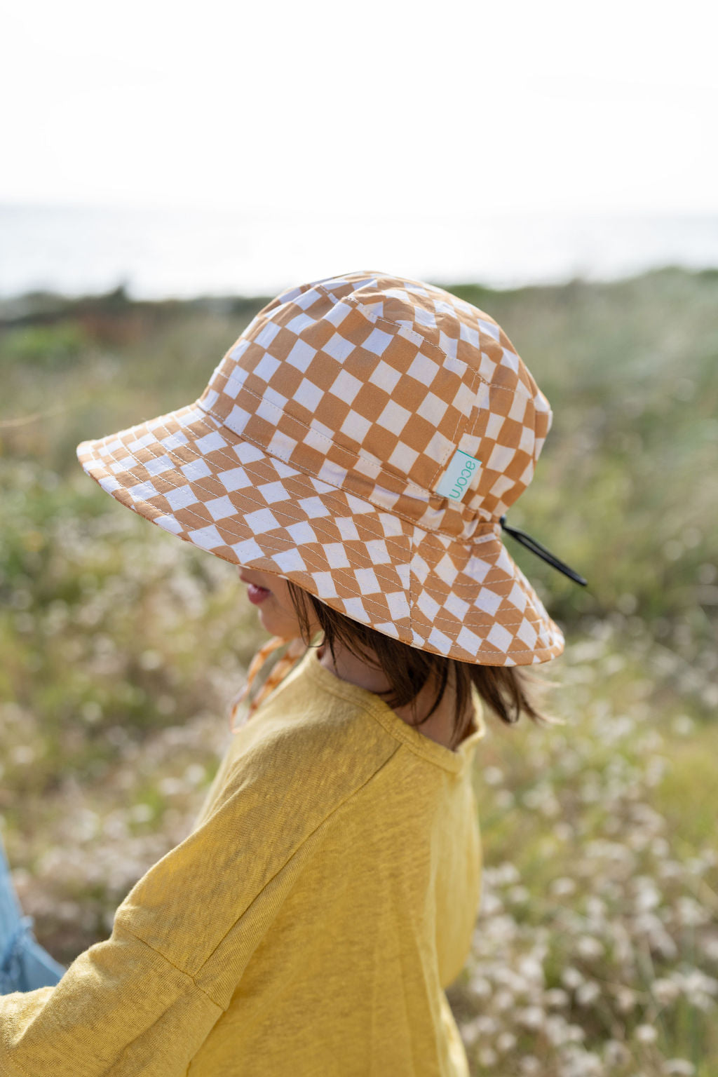 A child in a yellow shirt is outdoors, sporting the ACORN KIDS Checkmate Wide Brim Bucket Hat featuring caramel and cream checks. The bright sun creates a natural setting with blurred greenery in the background. The child, protected by UPF50+ sun protection, looks downwards with a relaxed posture.