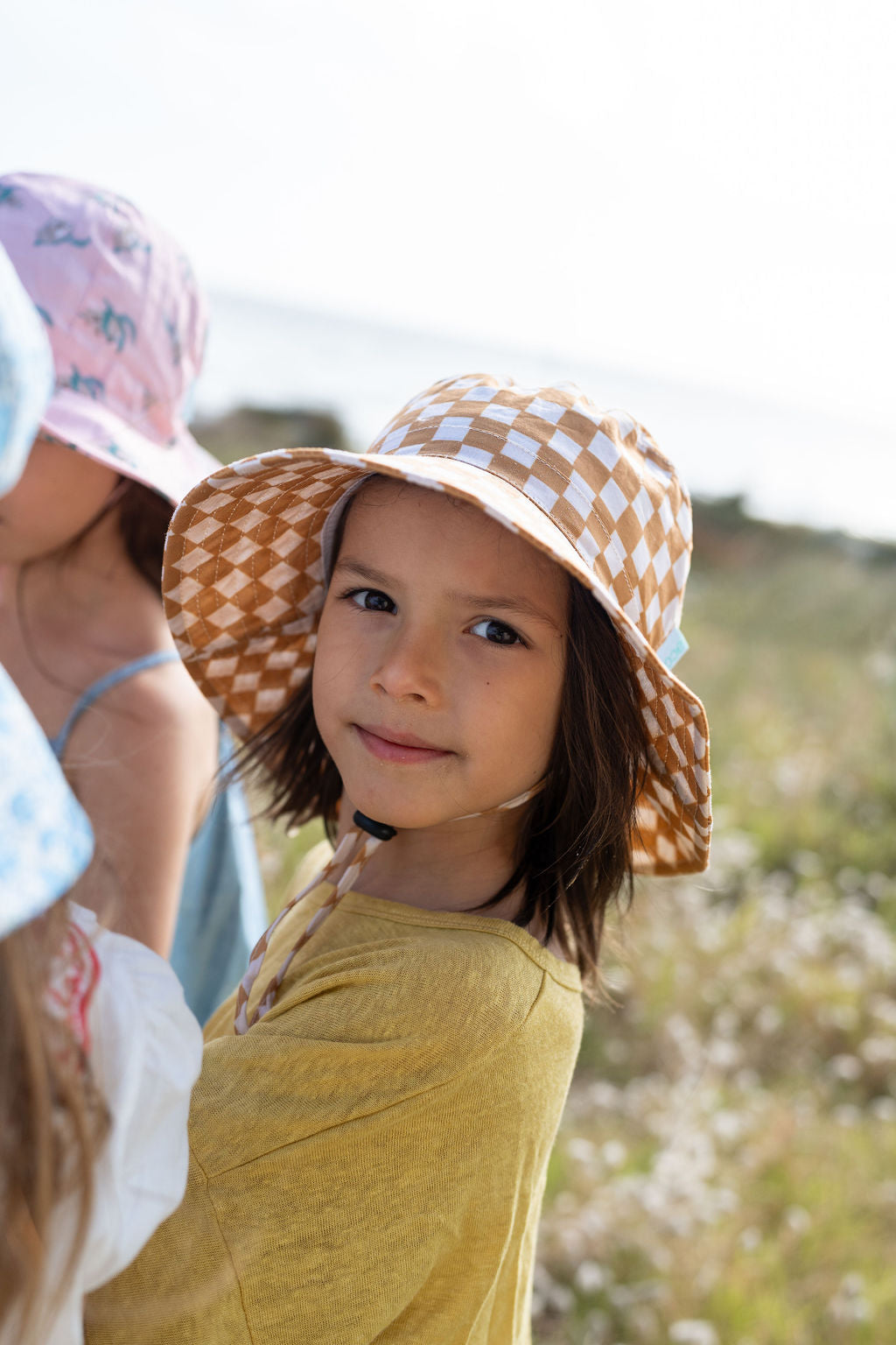 A child wearing an ACORN KIDS Checkmate Wide Brim Bucket Hat in beige checkered print with UPF50+ sun protection and a yellow top stands outside, smiling softly at the camera. The background shows another child with a floral hat and a blurred, sunny outdoor setting with greenery.