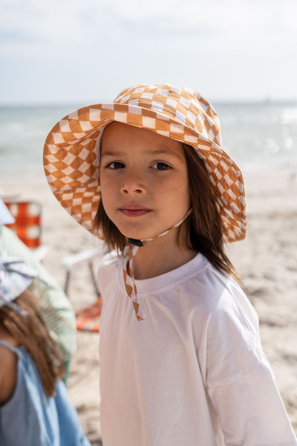 A young child with long hair is wearing a white shirt and the adorable ACORN KIDS Checkmate Wide Brim Bucket Hat in caramel and cream checks. They are standing on a sandy beach with the ocean in the background, looking directly at the camera.