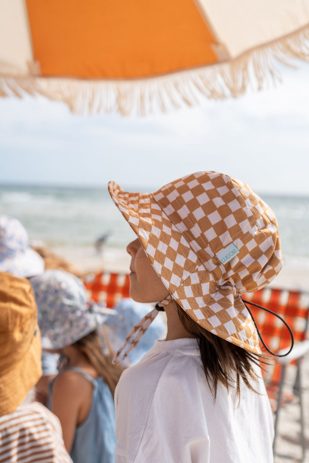 A child wearing the Checkmate Wide Brim Bucket Hat by ACORN KIDS stands under a fringed beach umbrella, gazing towards the sea. Other children in hats and summery clothes with UPF50+ sun protection are visible in the background, enjoying the beach scenery. The sky is clear and the ocean waves are gentle.