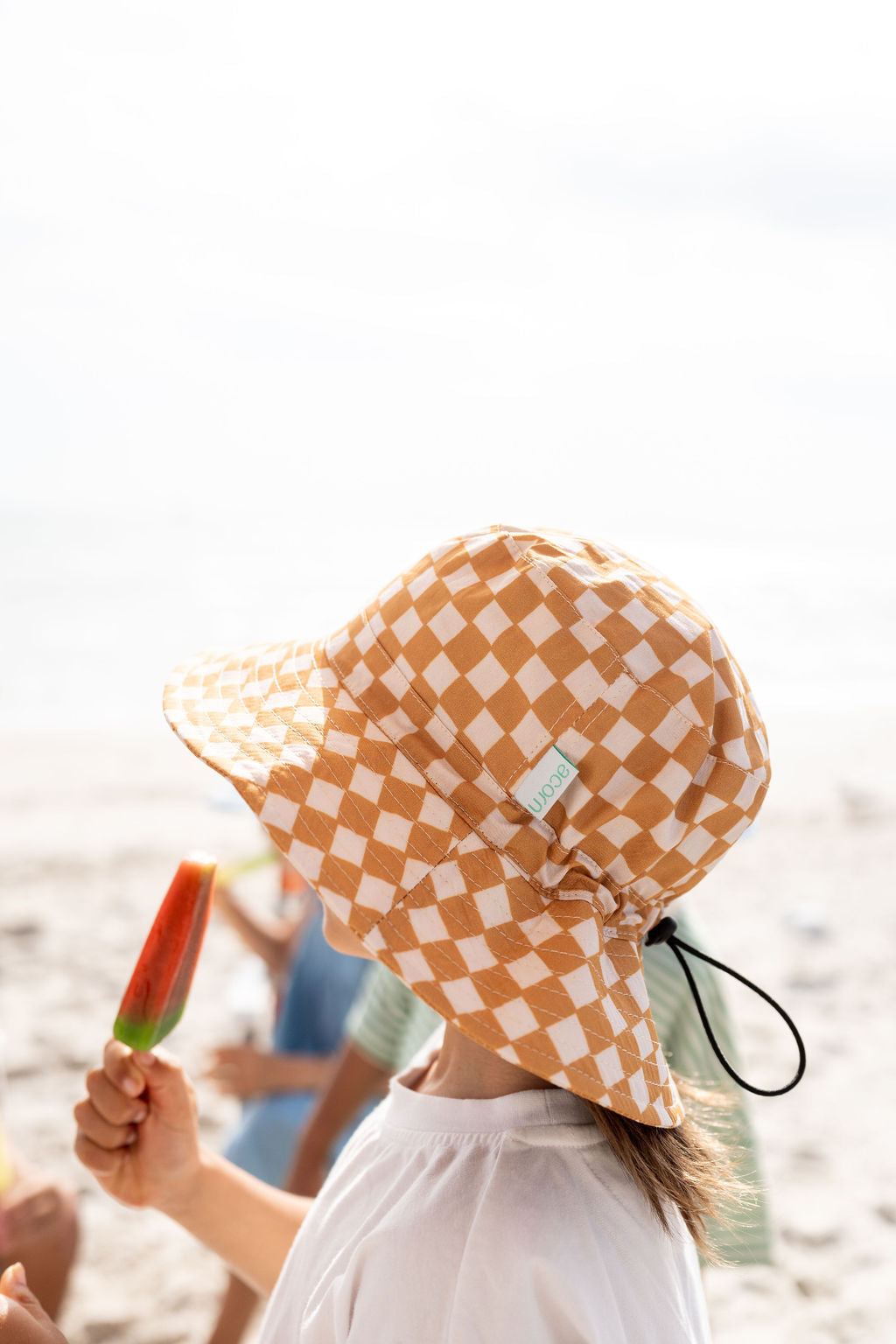 A person wearing the ACORN KIDS Checkmate Wide Brim Bucket Hat and a white shirt holds an orange popsicle. The background reveals a blurry beach scene with bright sunlight, indicating a warm, sunny day.