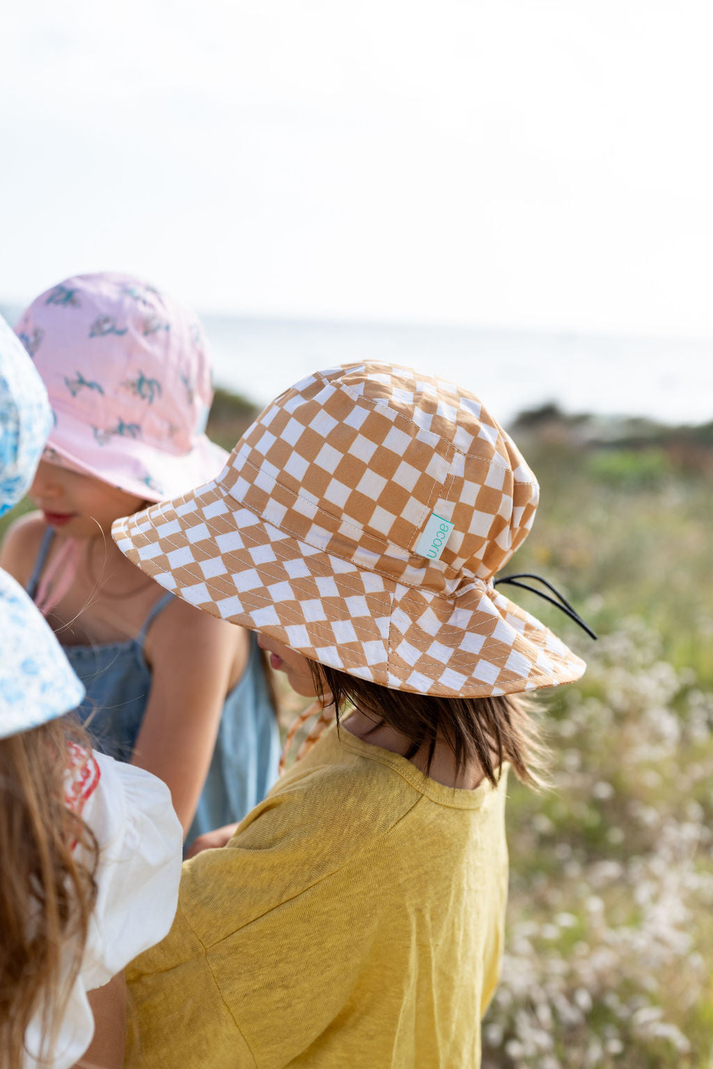 A group of children wearing sun hats stand outside on a sunny day. The child in focus wears a yellow shirt and an ACORN KIDS Checkmate Wide Brim Bucket Hat with caramel and cream checks, boasting UPF50+ sun protection. Other children, wearing pink and blue hats, are partially visible. Natural scenery with plants and a cloudy sky is in the background.