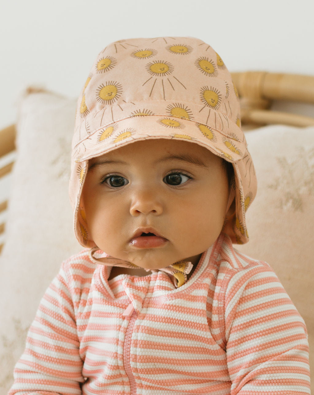A baby in the ACORN KIDS Sunshine Flap Hat—a light pink UPF50+ cap with yellow floral print—plus a pink and white striped onesie, sits on a pale cushion and gazes off to the side.