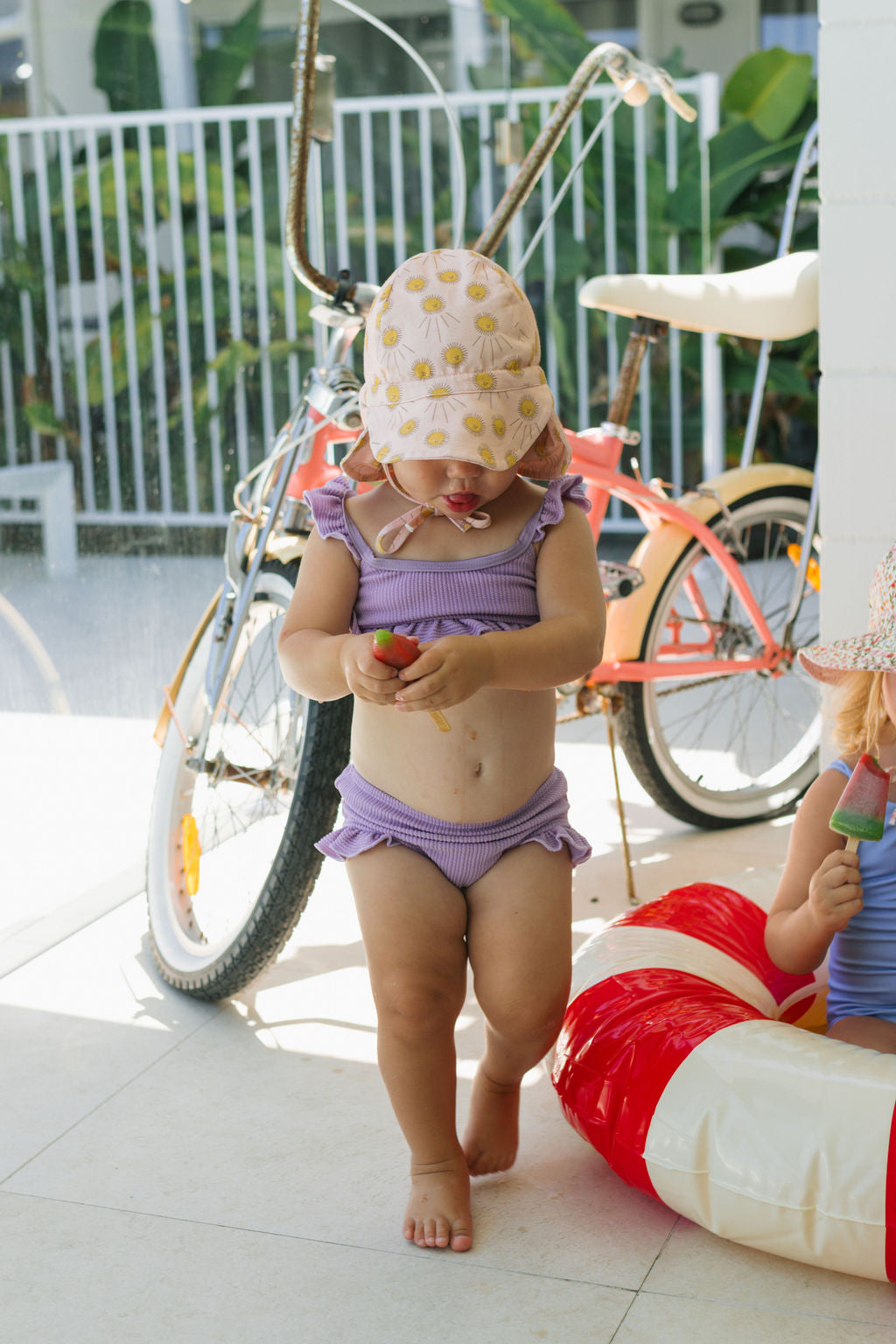 A toddler in a purple ruffled swimsuit and the ACORN KIDS Acorn Kids - Sunshine Flap Hat (UPF50+) walks indoors holding a popsicle, with a red bicycle and inflatable pool toy in the background.