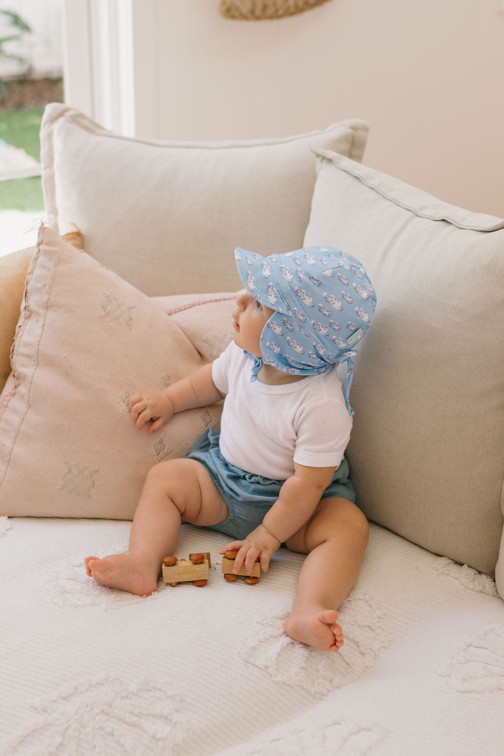 A baby in an ACORN KIDS Acorn Kids - Clouds Flap Hat and blue shorts sits on a couch, holding a wooden toy car. The soft cotton jersey hat offers UPF50+ sun protection. The couch features light-colored pillows and a blanket.
