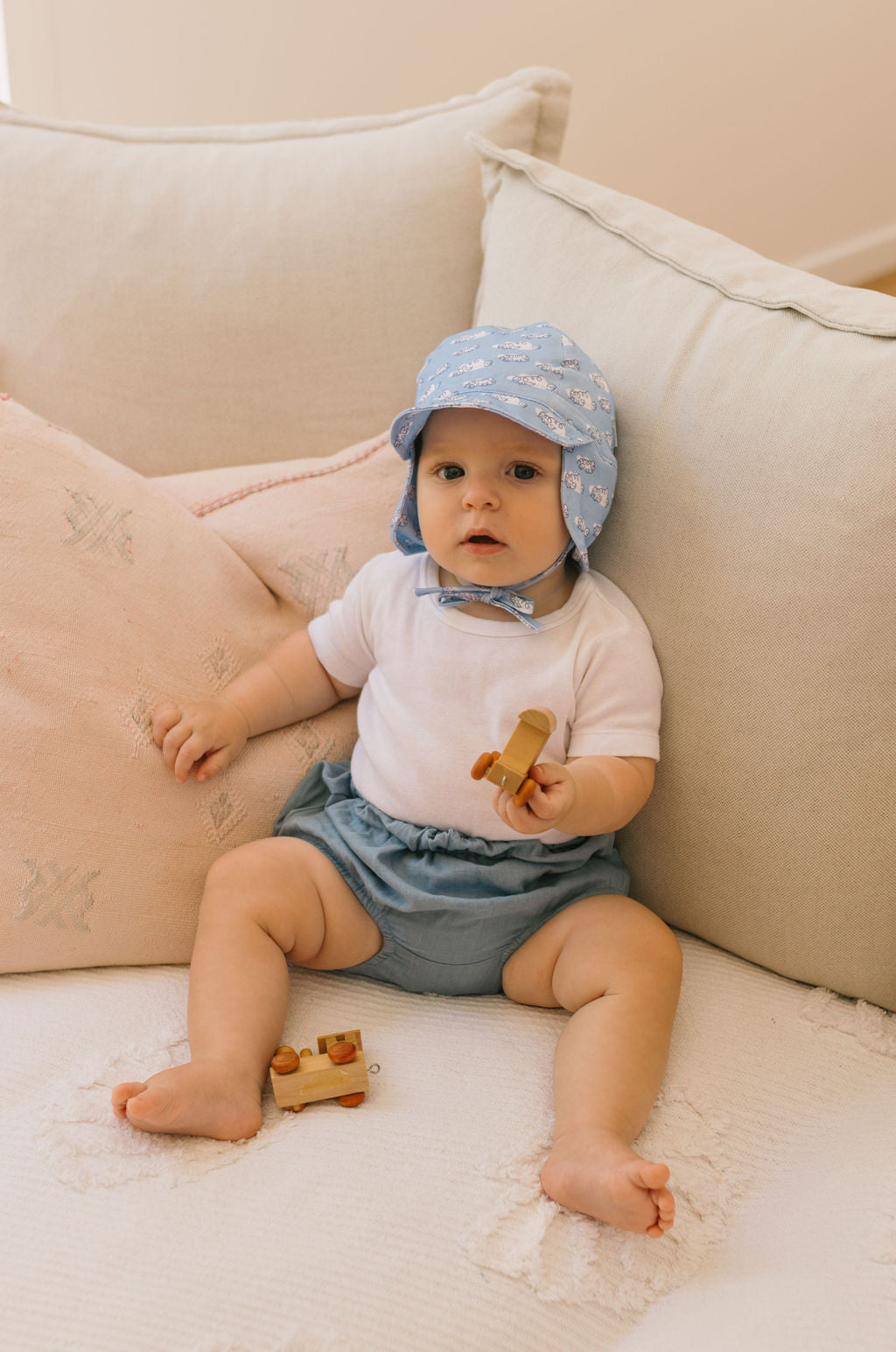 A baby wearing the ACORN KIDS Acorn Kids - Clouds Flap Hat with UPF50+ sun protection sits on a cream couch in a white shirt and blue shorts, holding a small wooden toy airplane with another toy by their legs.