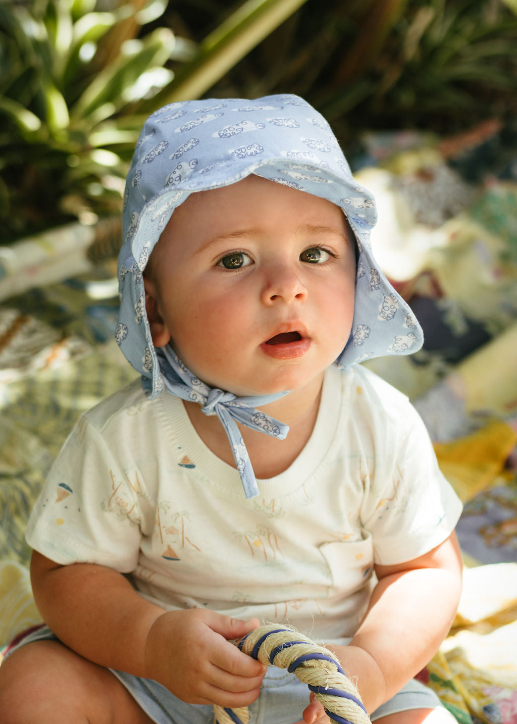 A baby in an ACORN KIDS Acorn Kids - Clouds Flap Hat with UPF50+ sun protection and a white shirt sits outside on a patterned blanket, holding a rope, surrounded by sunlight and greenery.
