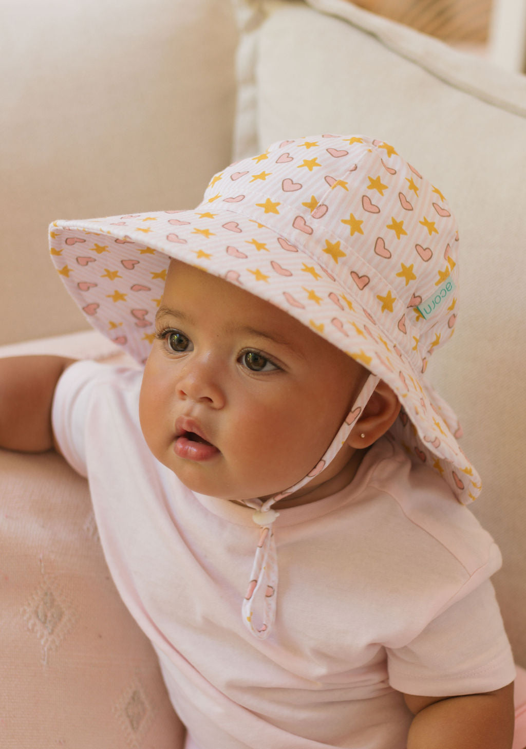 A baby wears the ACORN KIDS Acorn Kids - Love and Stars Stretch Cotton Wide Brim Sunhat, featuring a pink and yellow heart and star pattern, while sitting on a light-colored couch in a pale pink cotton jersey shirt.