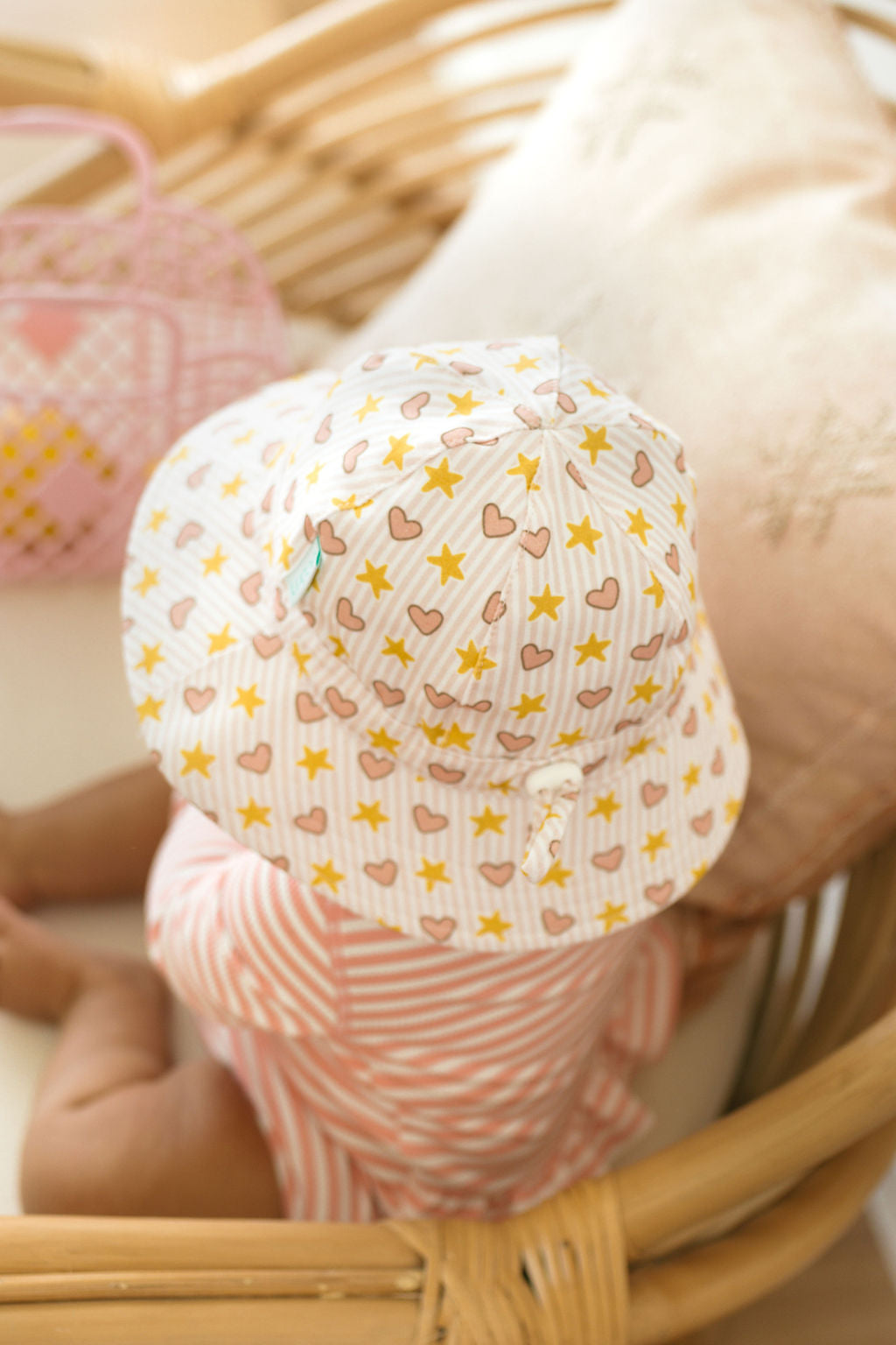 A baby in an ACORN KIDS Love and Stars Stretch Cotton Wide Brim Sunhat sits in a wicker bassinet, dressed in a pink and white striped outfit. A pillow and a small bag are nearby.