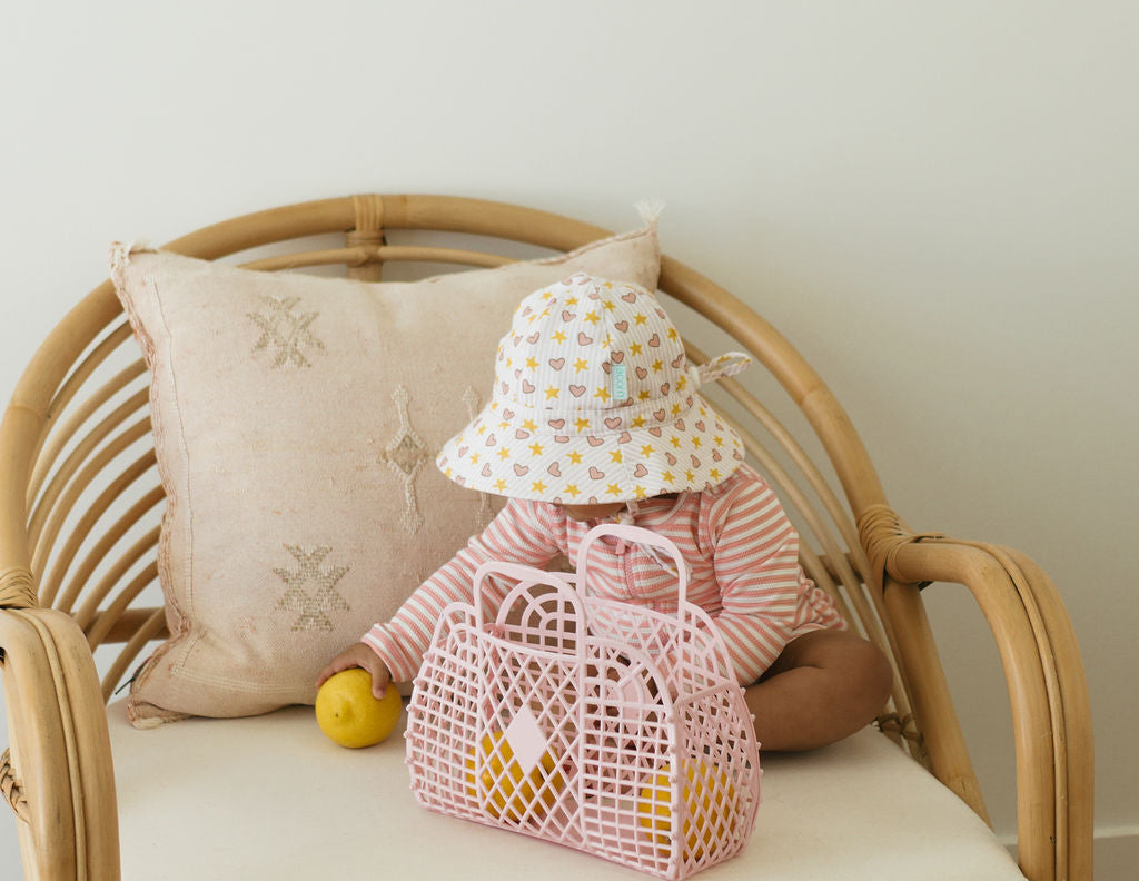 A baby in the ACORN KIDS Love and Stars Stretch Cotton Wide Brim Sunhat sits on a rattan chair, wearing a striped outfit, holding a yellow toy lemon next to a pink woven bag, with a beige cushion for comfort.