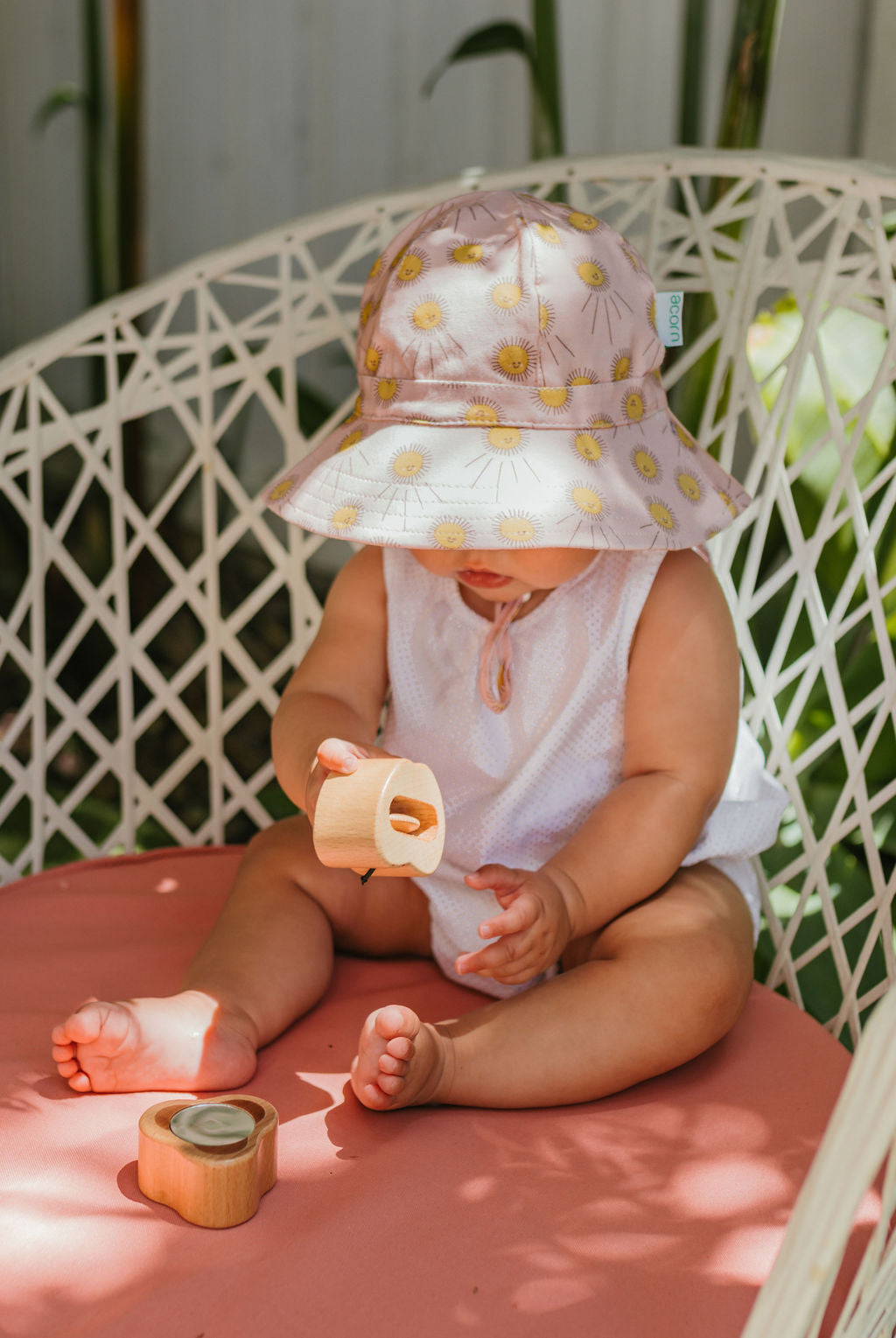 A baby in an ACORN KIDS Acorn Kids - Sunshine Stretch Cotton Wide Brim Sunhat and white outfit sits on a pink cushion in a woven chair, playing with wooden toys. The wide-brim sunhat partially covers the baby's face, with green plants in the background.