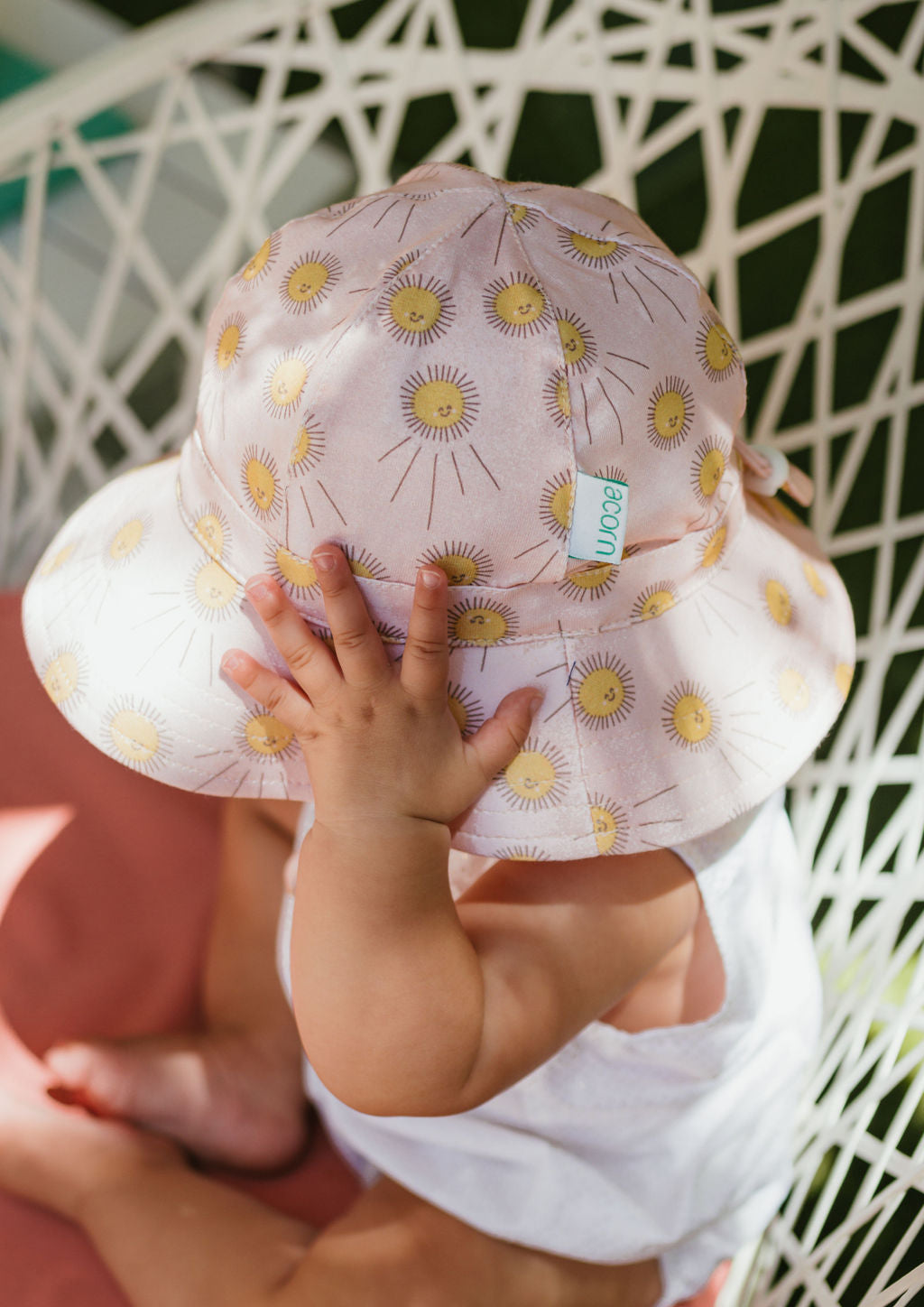 A baby in a white outfit sits in a white woven chair, touching the brim of their Acorn Kids - Sunshine Stretch Cotton Wide Brim Sunhat by ACORN KIDS. Their face is not visible.