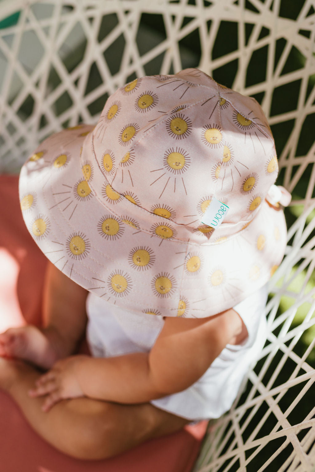 A baby in a white outfit and the ACORN KIDS Acorn Kids - Sunshine Stretch Cotton Wide Brim Sunhat sits on a pink cushion in a white woven chair, viewed from above.