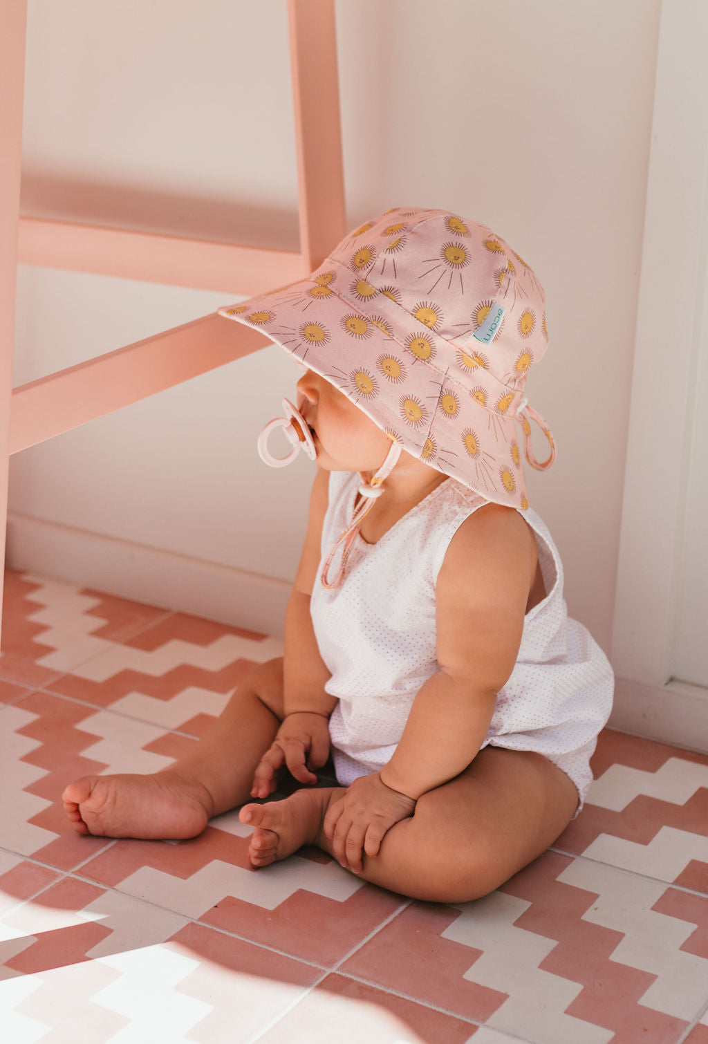 A baby in a white outfit and the ACORN KIDS Acorn Kids - Sunshine Stretch Cotton Wide Brim Sunhat sits on a pink and white patterned floor, looking to the side with a pacifier in their mouth.