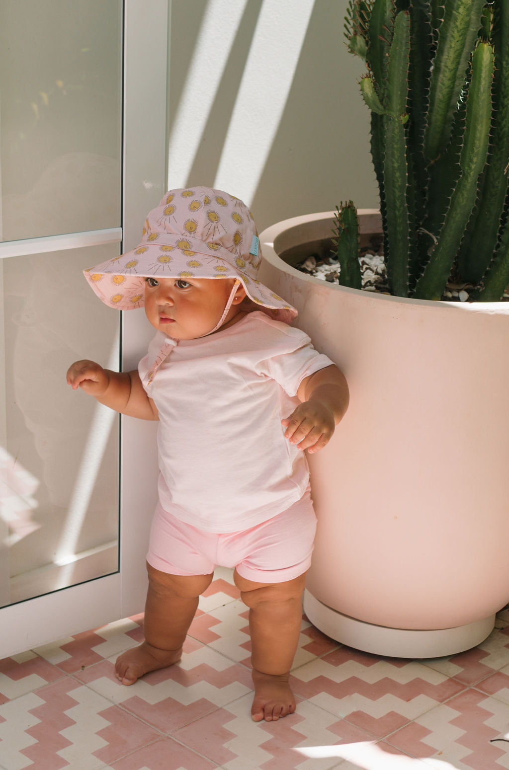A baby in a pink outfit and peach print Acorn Kids - Sunshine Stretch Cotton Wide Brim Sunhat by ACORN KIDS stands beside a large cactus on patterned tiles, sunlight highlighting the hat’s comfy UPF50+ sun protection.