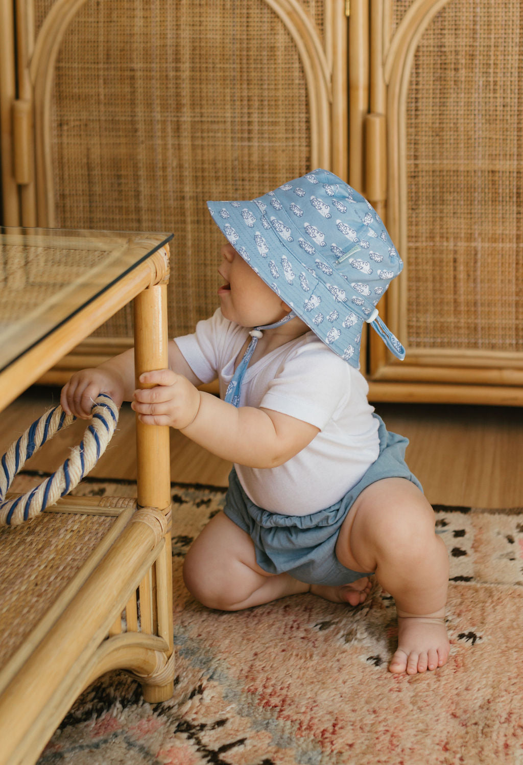 A baby in an Acorn Kids - Clouds Stretch Cotton Wide Brim Sunhat by ACORN KIDS and blue shorts kneels on a patterned rug, holding a bamboo and glass table, with wicker furniture behind. The UPF50+ sunhat offers excellent sun protection.