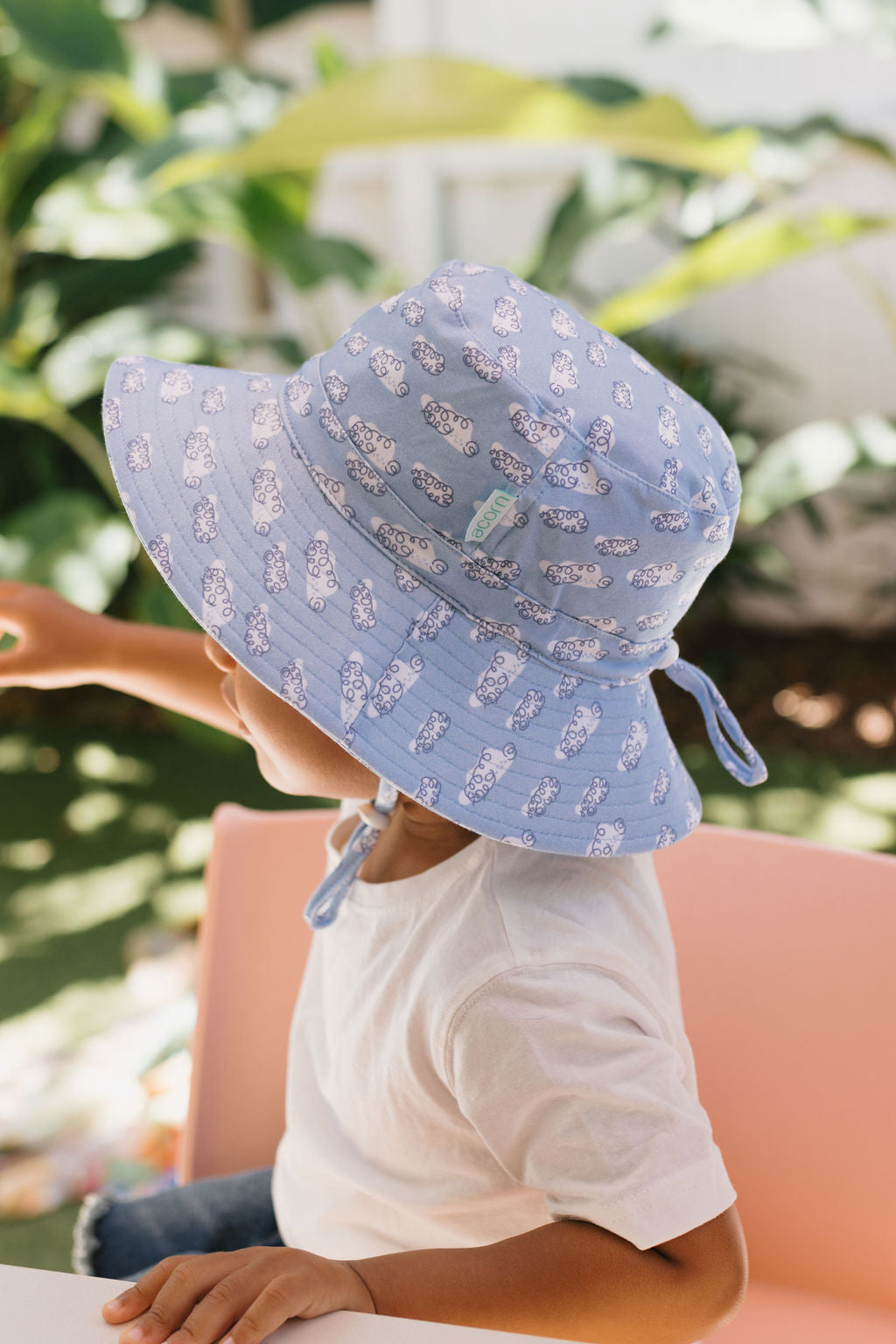 A child wears the Acorn Kids - Clouds Stretch Cotton Wide Brim Sunhat by ACORN KIDS while sitting on a pink chair outdoors, reaching out amid greenery in the background.