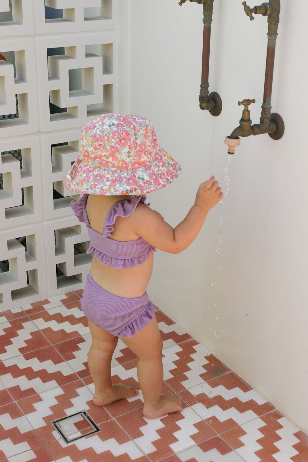 A toddler in a purple ruffled swimsuit and the ACORN KIDS Tessa Broad Brim Bucket Hat stands on red-and-white tiles, reaching for water from an outdoor faucet on a white wall.