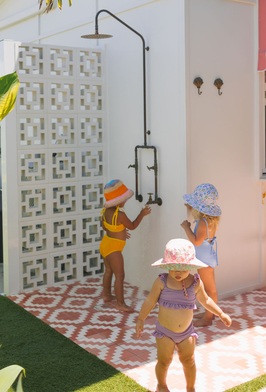 Three children wear ACORN KIDS’ Tessa Broad Brim Bucket Hats as they play near an outdoor shower with patterned tiles and a privacy screen. Sunlight casts shadows, but UPF50+ protection keeps them safe during their fun.