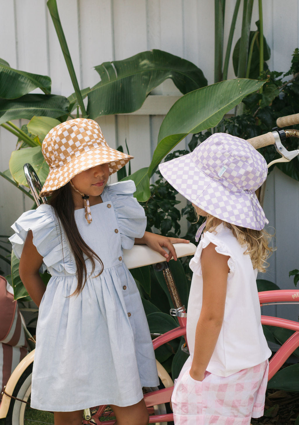 Two young girls stand by a pink bike wearing ACORN KIDS hats—one orange, one Acorn Kids Lavender Checks Broad Brim Bucket Hat with UPF50+ protection. One girl wears a light blue dress; the other wears a white top and pink checkered shorts. Green plants line a white fence behind them.