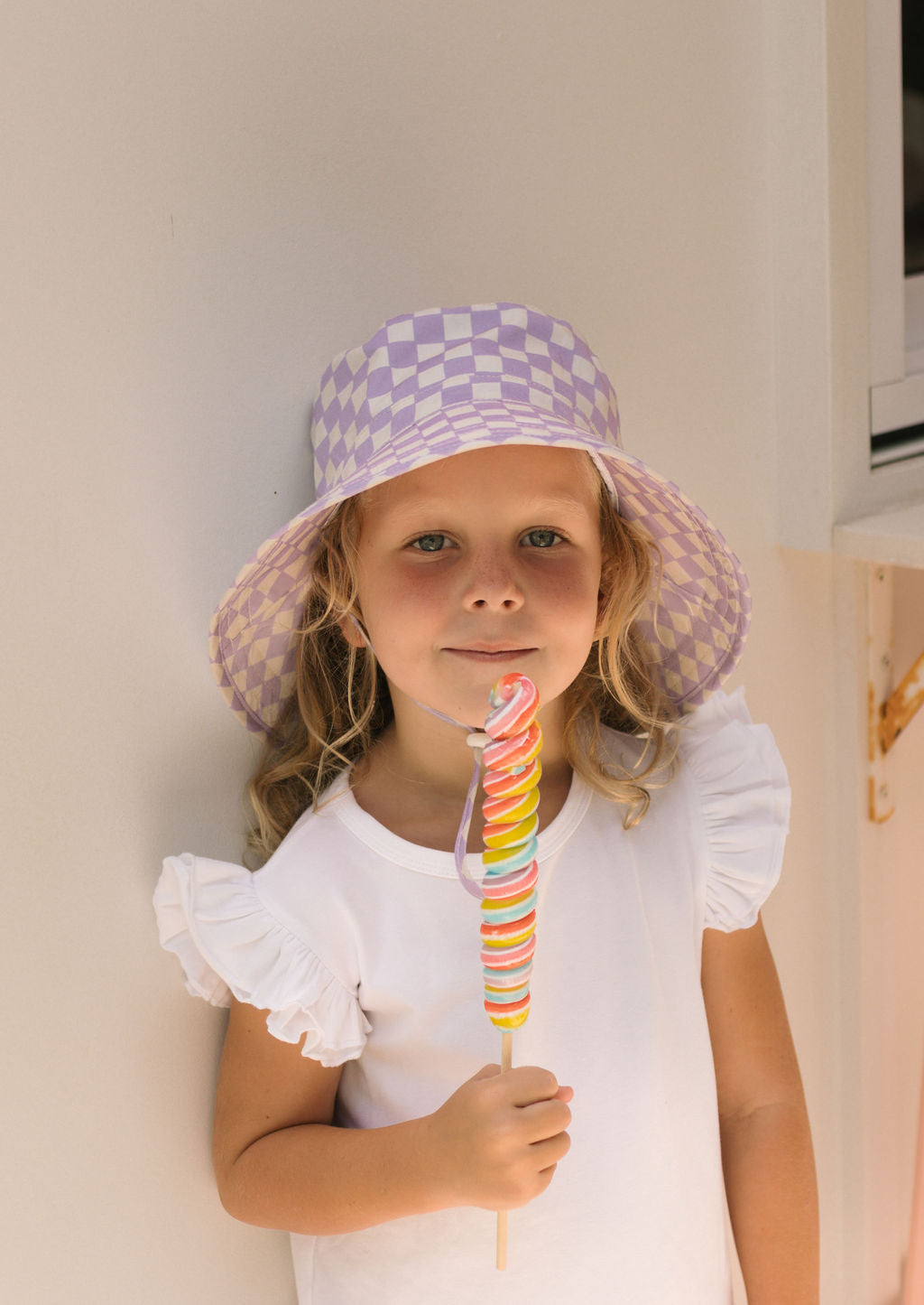 A young child with long blond hair wears the ACORN KIDS Acorn Kids - Lavender Checks Broad Brim Bucket Hat with UPF50+ sun protection and a white ruffled-sleeve shirt, standing against a light wall and holding a colorful spiral lollipop.