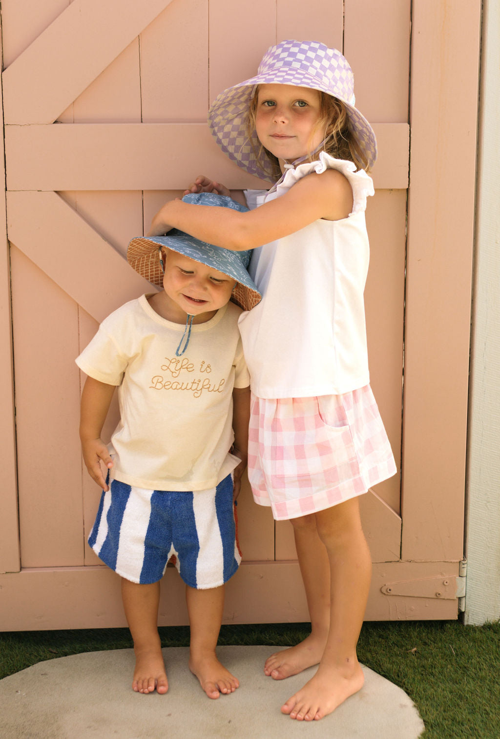 Two young children stand barefoot by a pink wooden gate. The older girl, in the Acorn Kids - Lavender Checks Broad Brim Bucket Hat by ACORN KIDS and shorts, hugs a younger boy wearing a blue bucket hat and striped shorts.