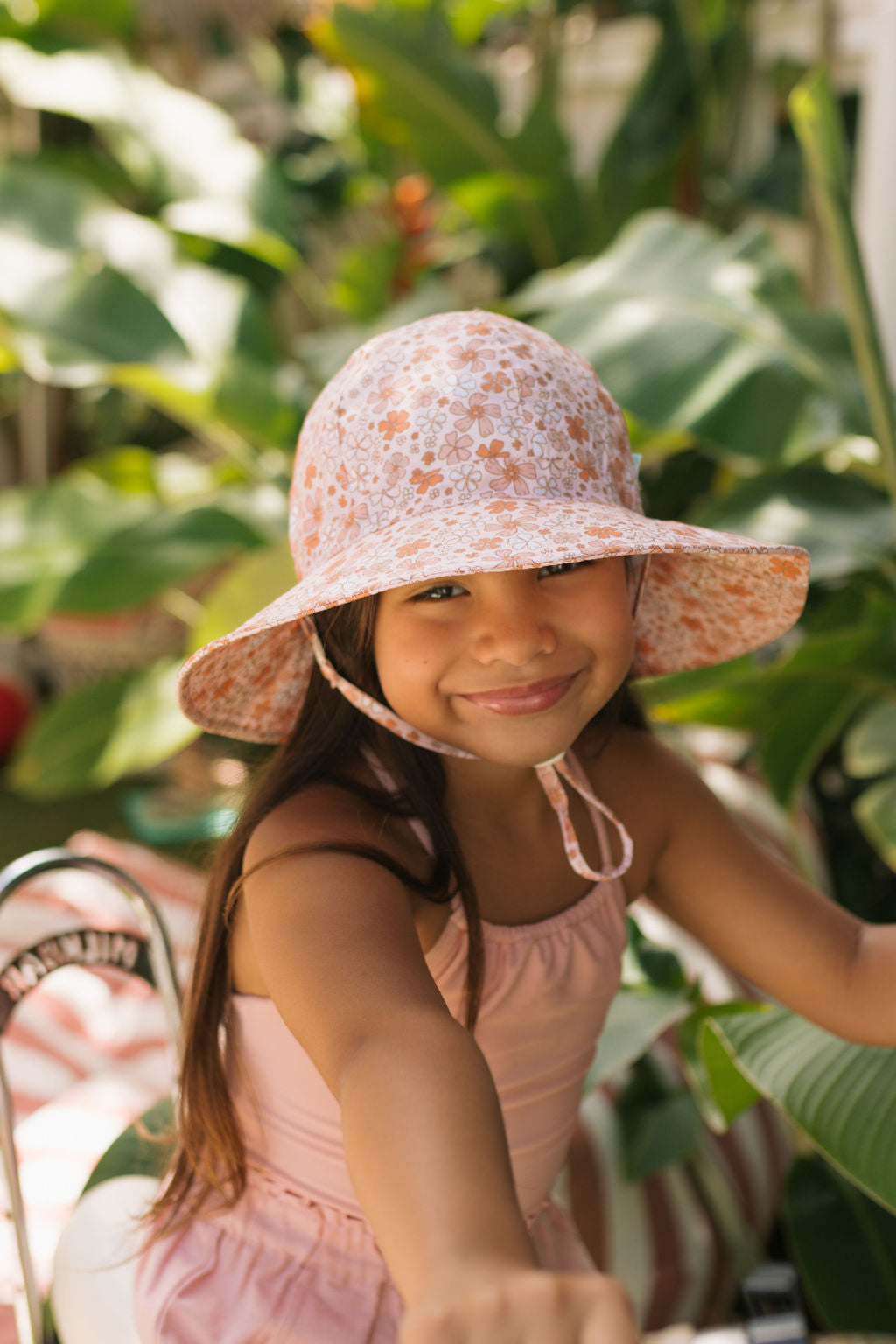 A young girl wearing the ACORN KIDS Acorn Kids - Lulu Wide Brim Sunhat and a light pink sleeveless outfit smiles and reaches forward while sitting outdoors among lush green plants.
