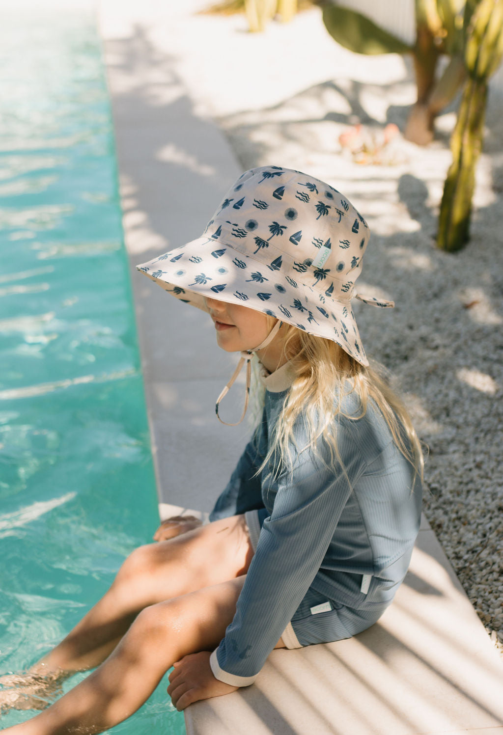 A young girl wearing the ACORN KIDS Acorn Kids - Island Breeze Wide Brim Bucket Hat and a long-sleeve swimsuit sits by the pool, feet in the water, relaxing in partial shade among gravel and lush green plants.