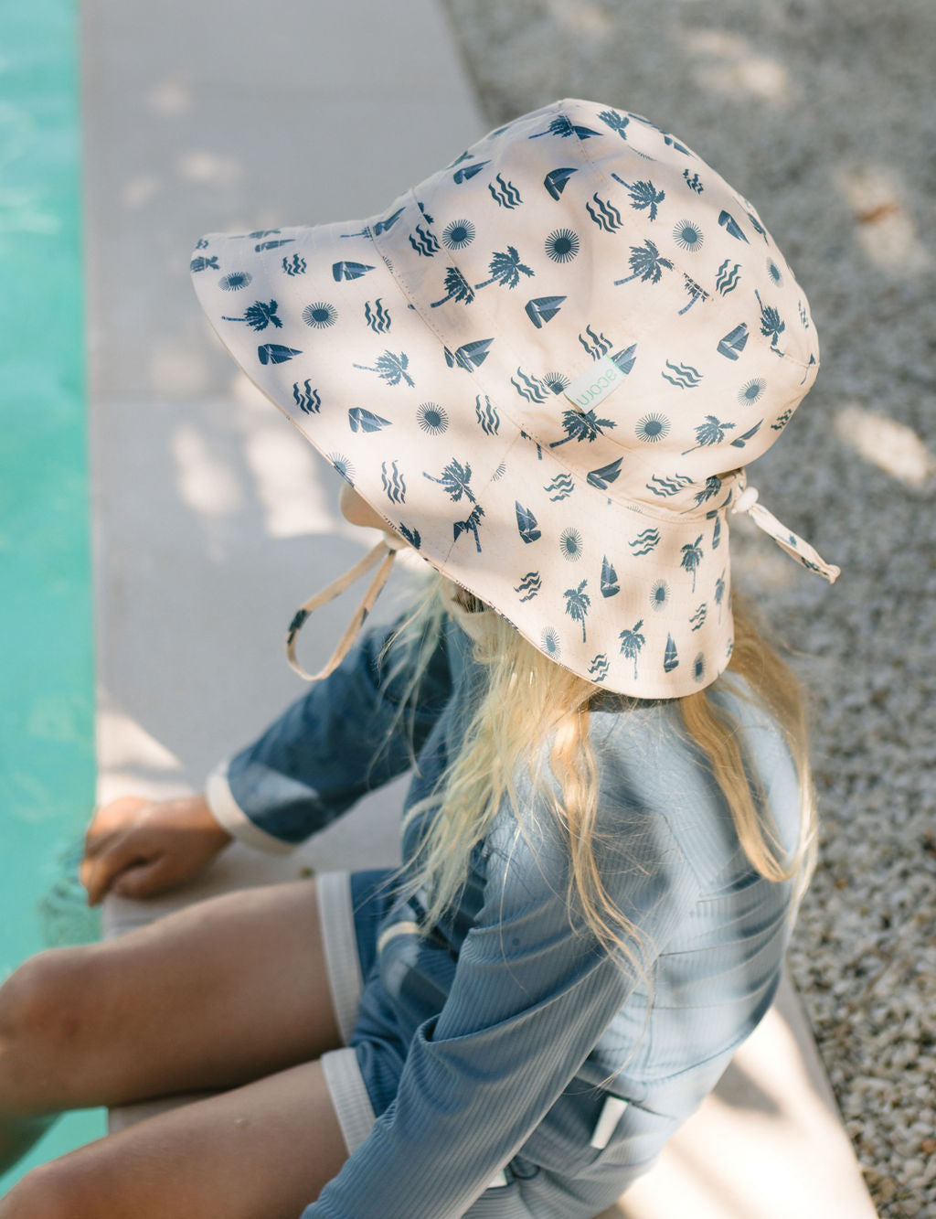 A person in a blue outfit sits by the pool, wearing the ACORN KIDS Acorn Kids - Island Breeze Wide Brim Bucket Hat, featuring blue nautical prints and UPF50+ sun protection as sunlight casts gentle shadows nearby.