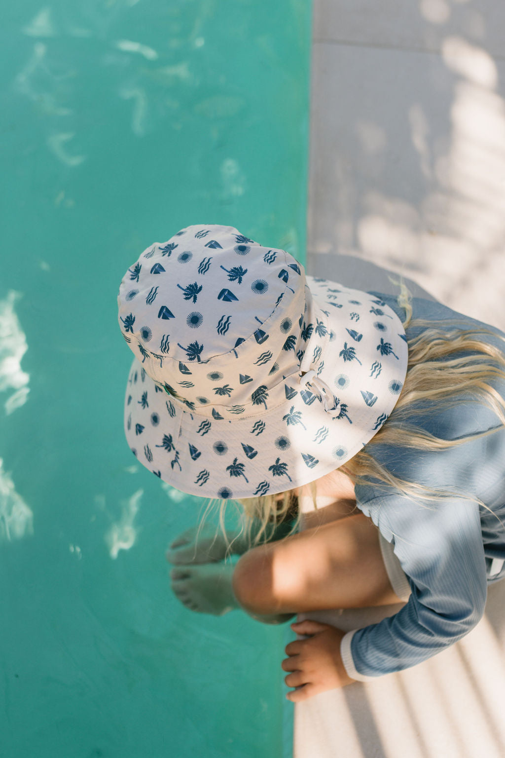 A child wearing the Acorn Kids - Island Breeze Wide Brim Bucket Hat by ACORN KIDS, featuring a blue palm tree pattern and UPF50+ sun protection, sits by the pool with toes in the water as sunlight casts gentle shadows.