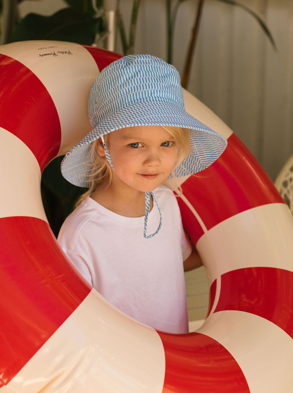A young child wearing the ACORN KIDS Acorn Kids - Mykonos Wide Brim Bucket Hat stands inside a large red and white inflatable pool ring, gently smiling at the camera in a white t-shirt.