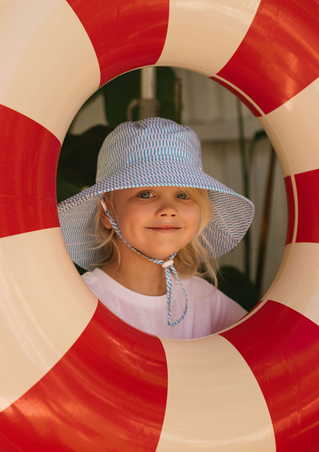 A smiling child in the ACORN KIDS Mykonos Wide Brim Bucket Hat looks through a red and white striped lifebuoy, staying cool and protected with 100% cotton fabric and UPF50+ sun protection.