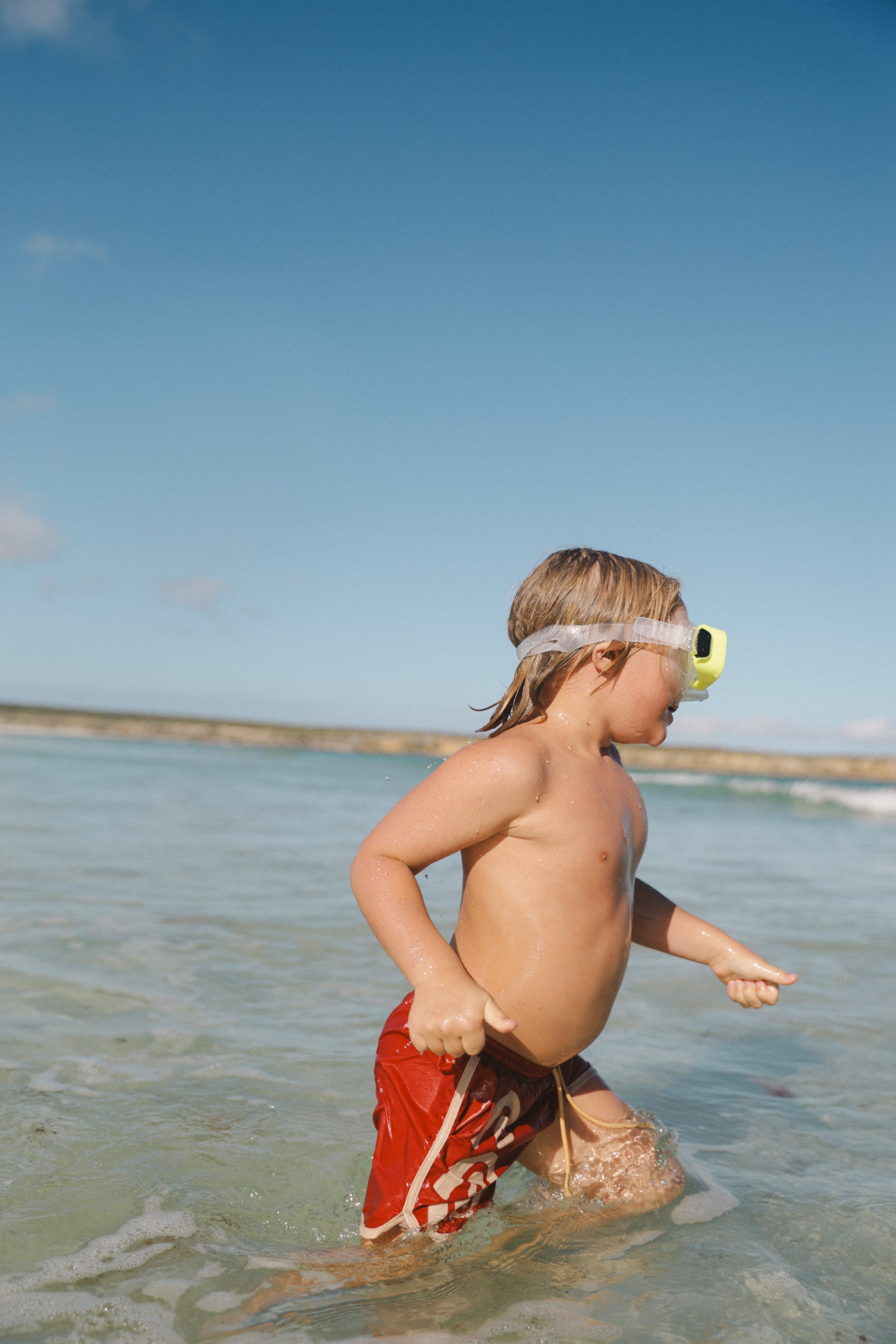 A child with long hair, wearing goggles and SMALL SWIM CLUB's Maroon Swirl Boardshorts featuring an elastic drawstring waistband, plays in the shallow ocean under a sunny, clear blue sky.