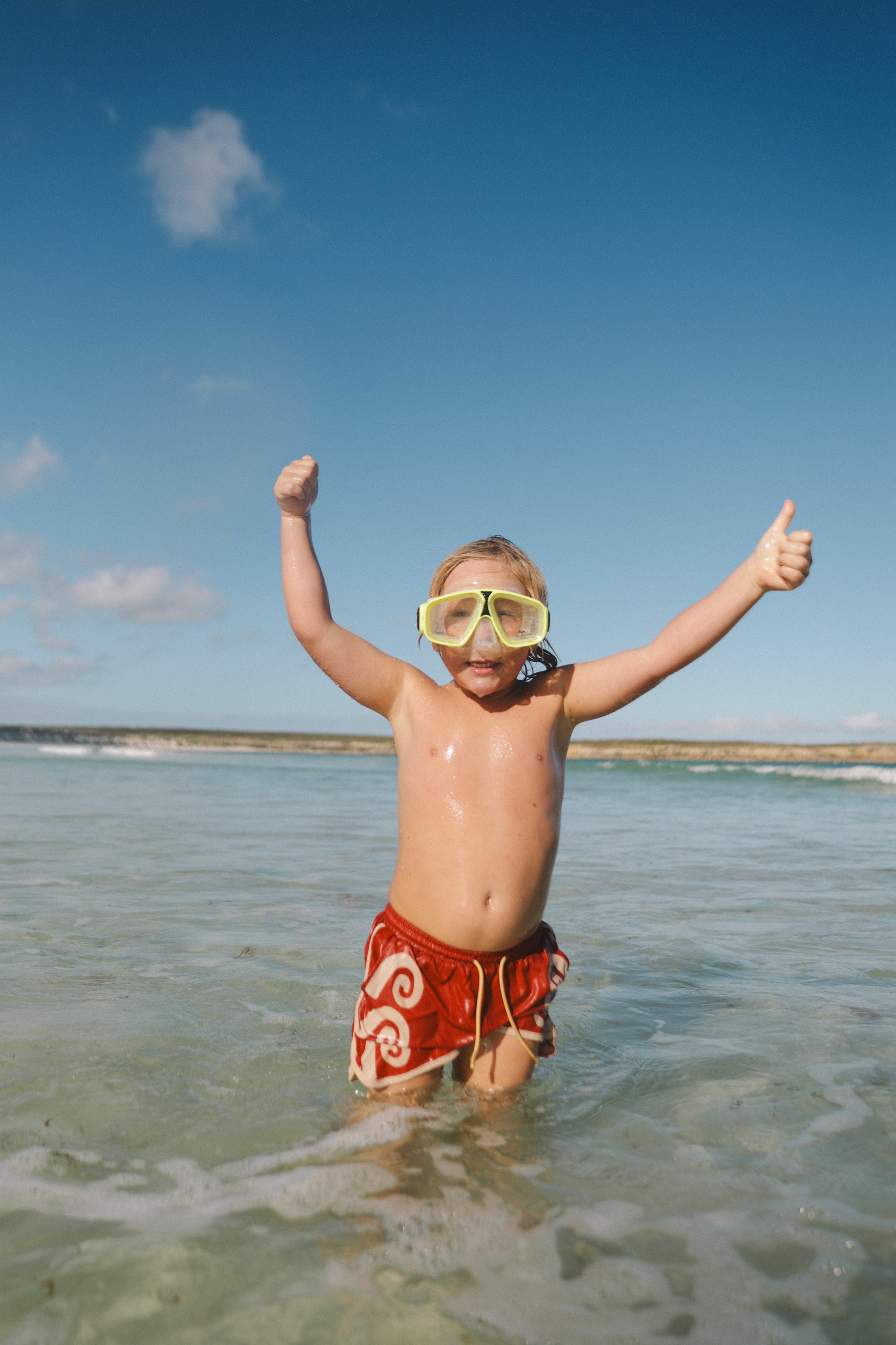 A child in SMALL SWIM CLUB's Maroon Swirl Boardshorts with an elastic drawstring waistband and yellow diving goggles stands in shallow water, arms raised and giving two thumbs up. A clear sky and calm ocean set the perfect backdrop.