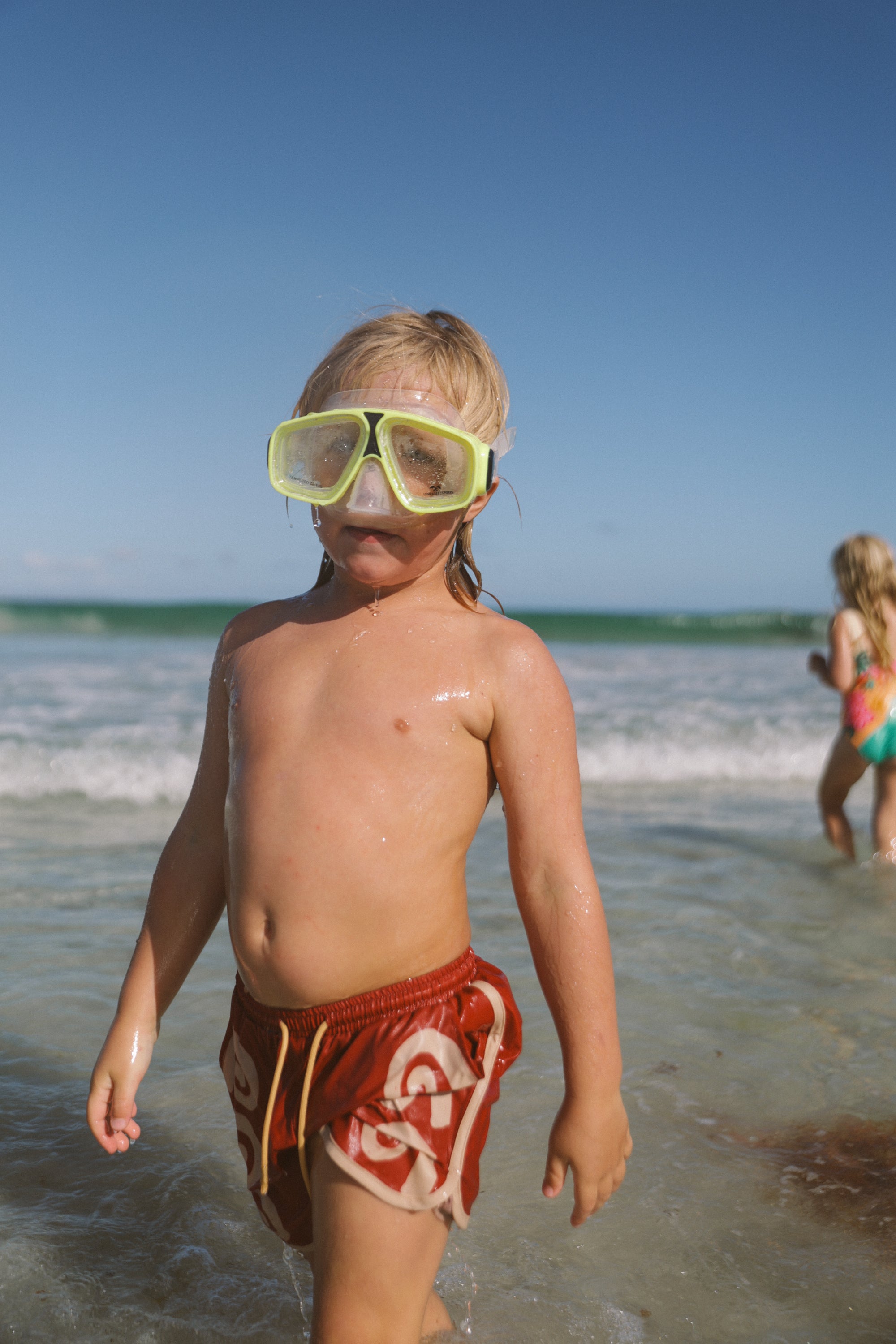 A young child in SMALL SWIM CLUB's Maroon Swirl Boardshorts with an elastic waistband and yellow goggles stands at the ocean's edge, wet from a recent swim, while another child plays near the waves under a clear blue sky.