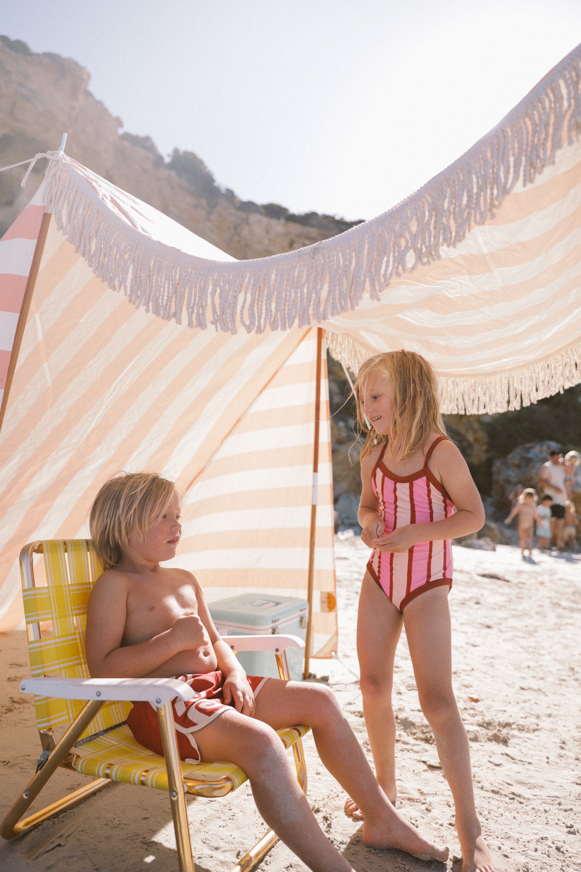 Two kids under a striped beach canopy; one sits in a yellow chair wearing SMALL SWIM CLUB's Maroon Swirl Boardshorts with an elastic drawstring, while the other stands in a red and white striped swimsuit. Sandy beach and rocky cliffs are in the background.