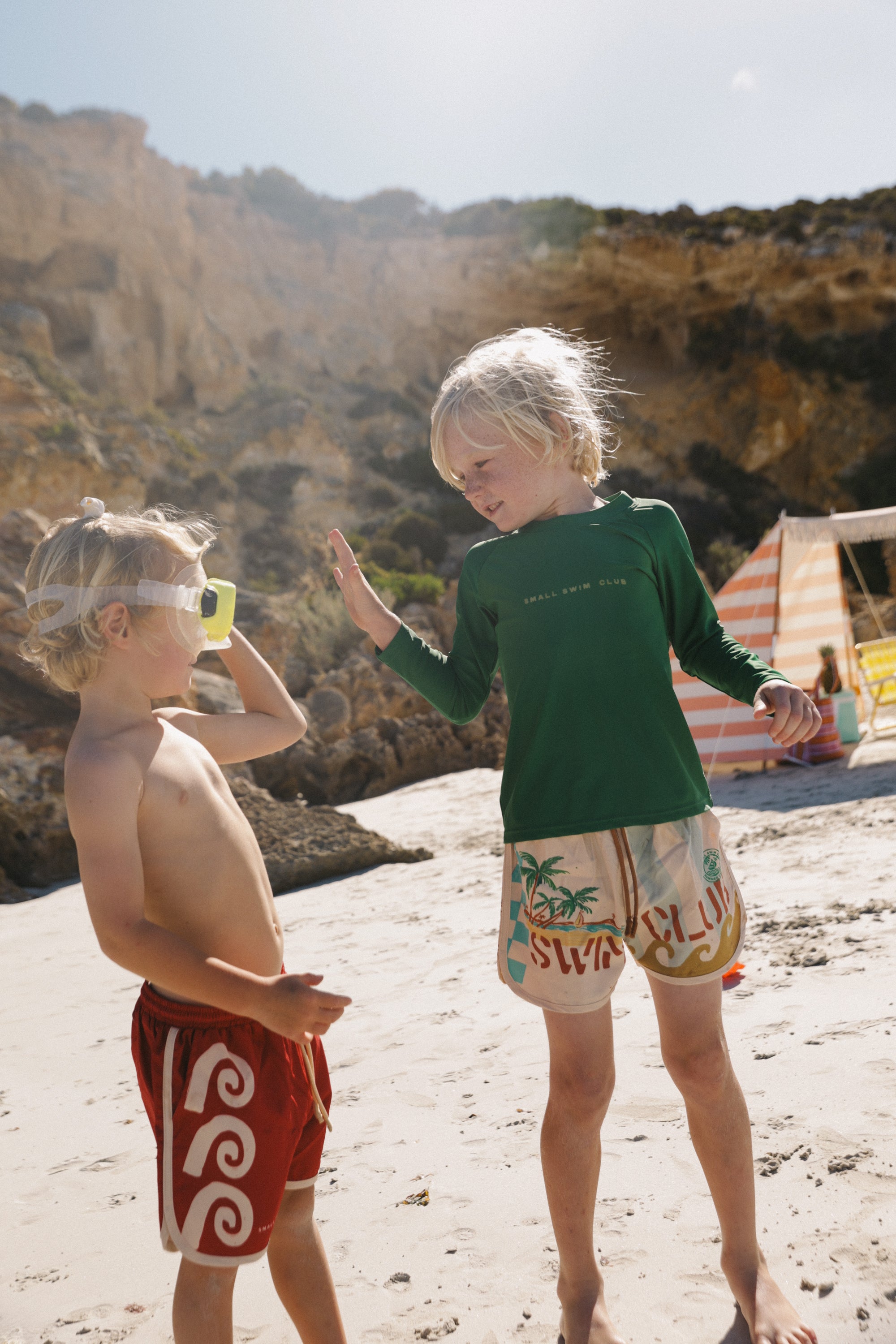 Two children stand on a sandy beach with rocky cliffs behind them. One in red shorts and goggles, the other raises a hand wearing a green shirt and SMALL SWIM CLUB's Maroon Swirl Boardshorts with an elastic drawstring waistband. A striped beach tent is visible in the background.