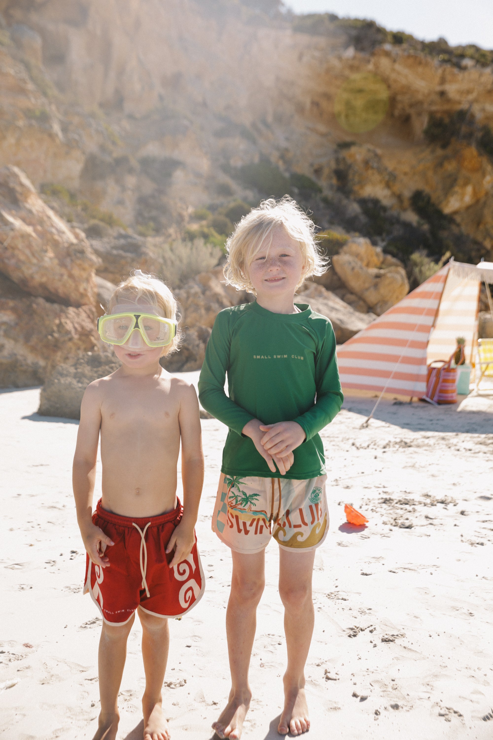 On a sunny beach, two kids in SMALL SWIM CLUB trunks stand together. One wears yellow goggles and red trunks; the other sports Maroon Swirl Boardshorts. Behind them, a striped tent and rocky cliffs form the picturesque backdrop.